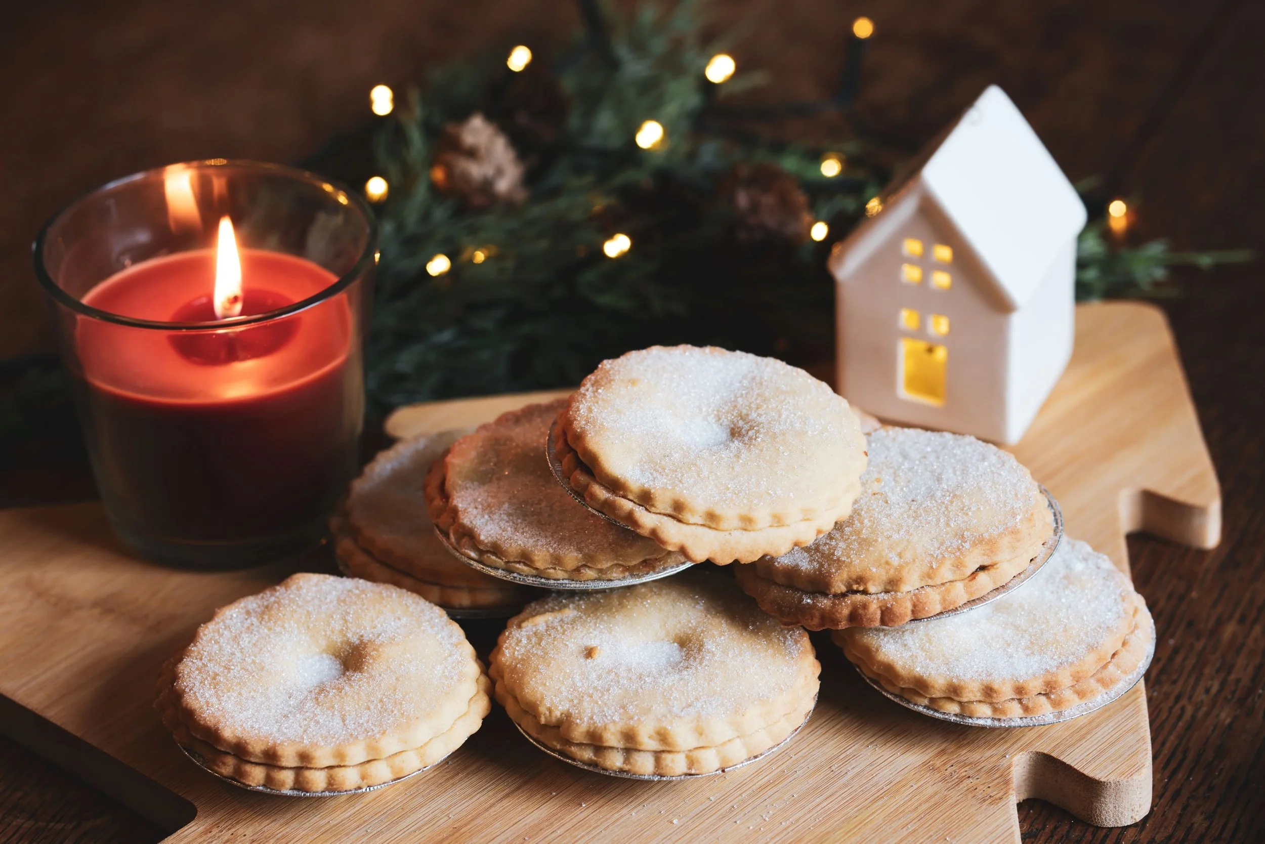 Cookies on a wooden tray with a lit red candle and a small white ceramic house, with holiday greenery and lights in the background.