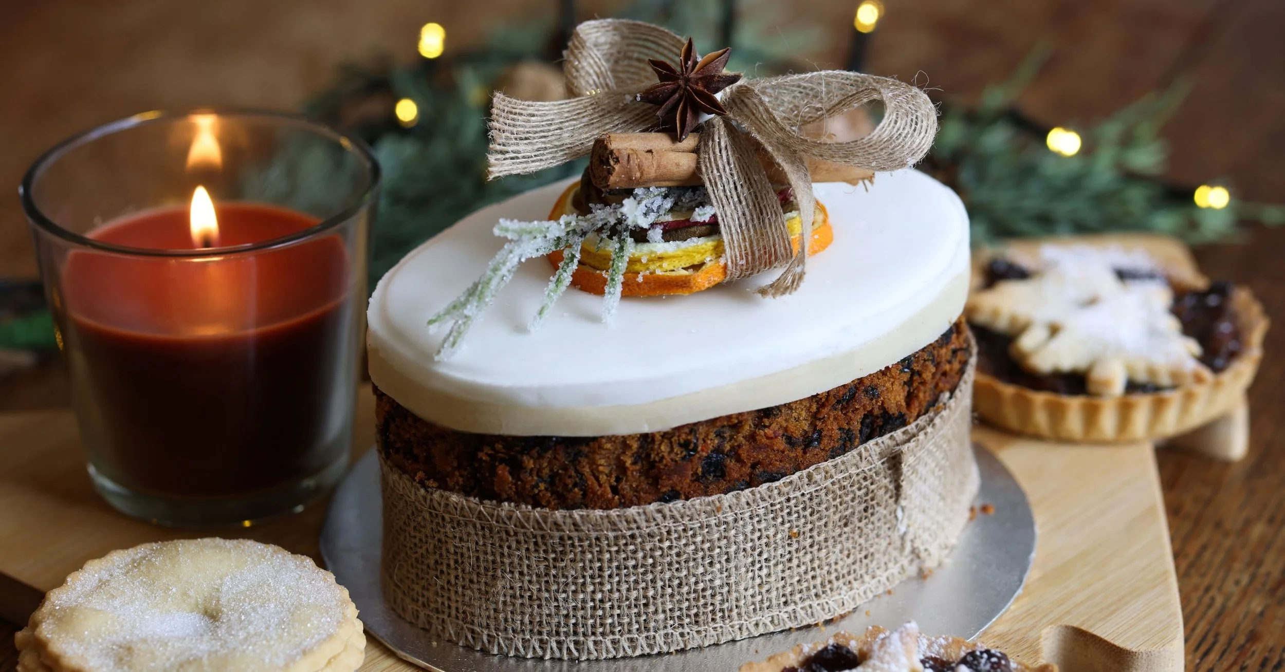 A Christmas-themed dessert table with a decorated cake, cookies, and a lit candle, surrounded by holiday greenery and lights.
