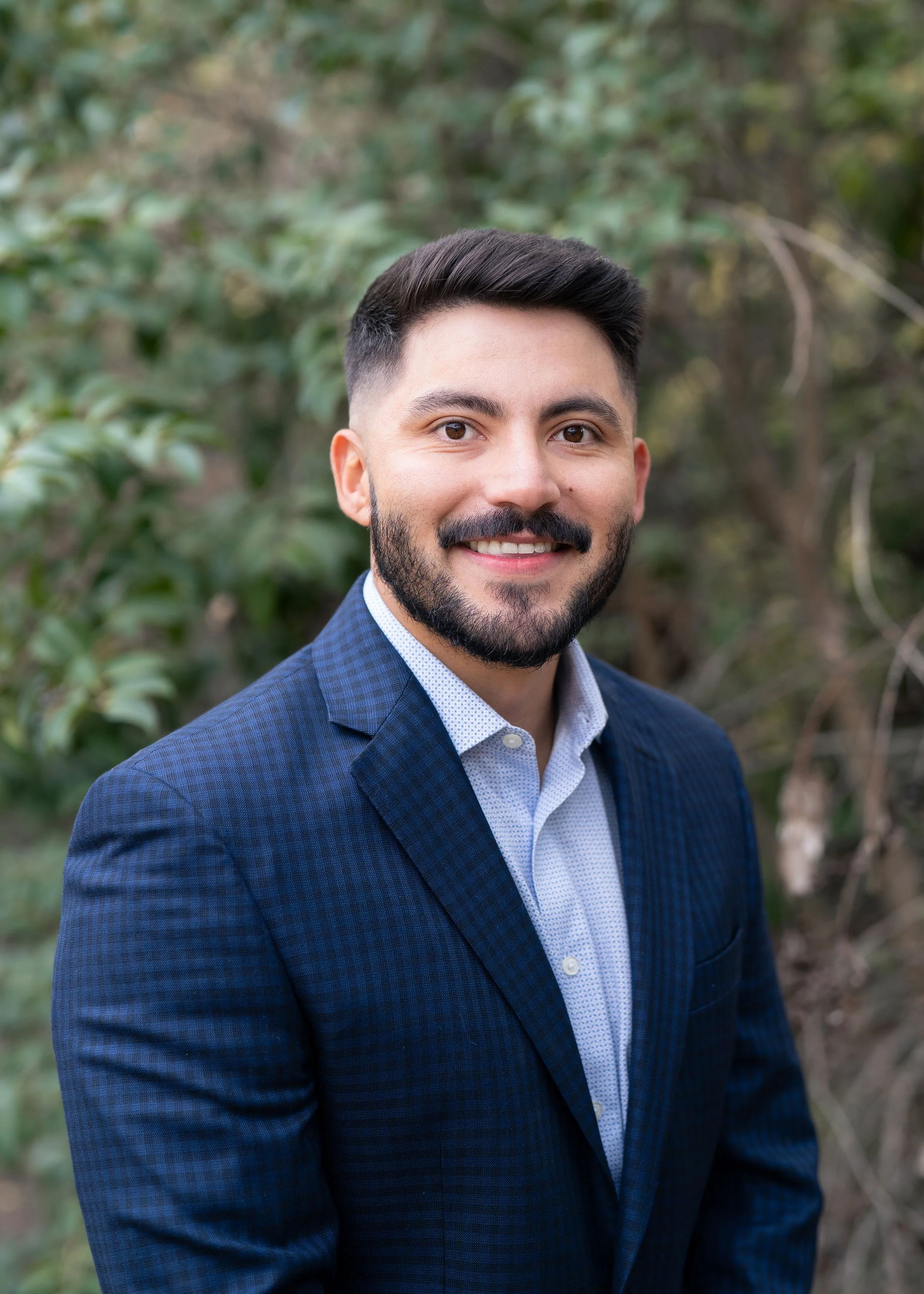 Black and white professional headshot of a young man with dark hair and beard, smiling, wearing a checkered blazer and white shirt, against a dark background.