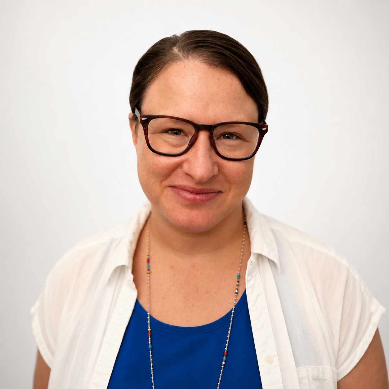 Close-up of a smiling woman with short brown hair, wearing red-framed glasses, a blue top, and a white shirt, standing against a plain light-colored wall.