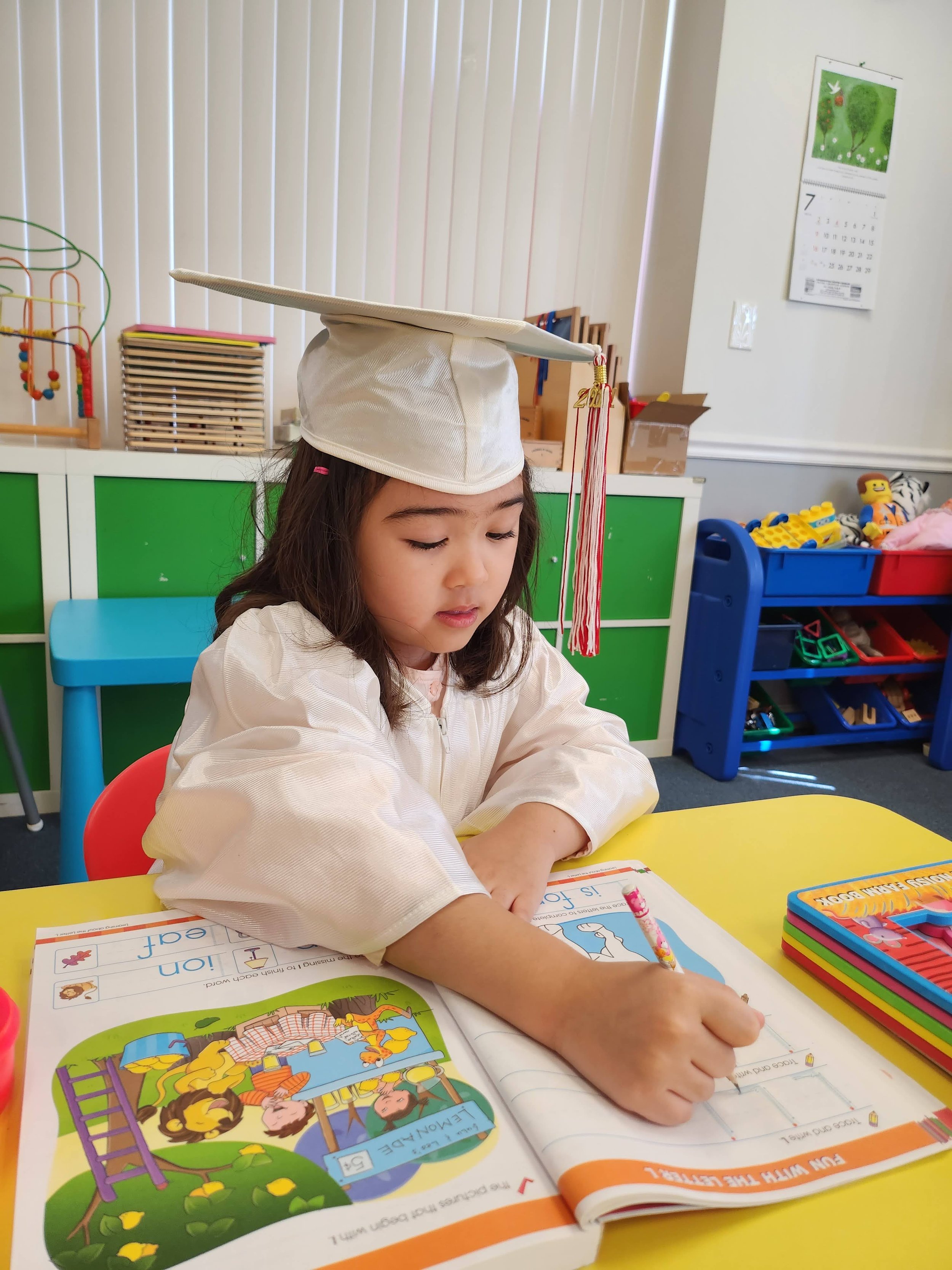 A young girl wearing a graduation cap and a white jacket is sitting at a yellow table, coloring on a workbook with a pink crayon. Behind her, there are colorful toy shelves, a green cabinet, a calendar on the wall, and a blue cart filled with toys.