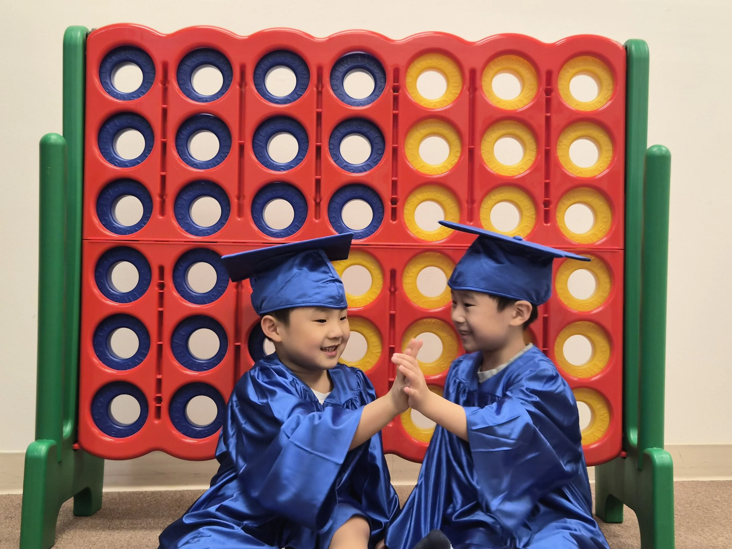 Two young boys wearing blue graduation gowns and caps, playing Connect 4 game together in an indoor setting.