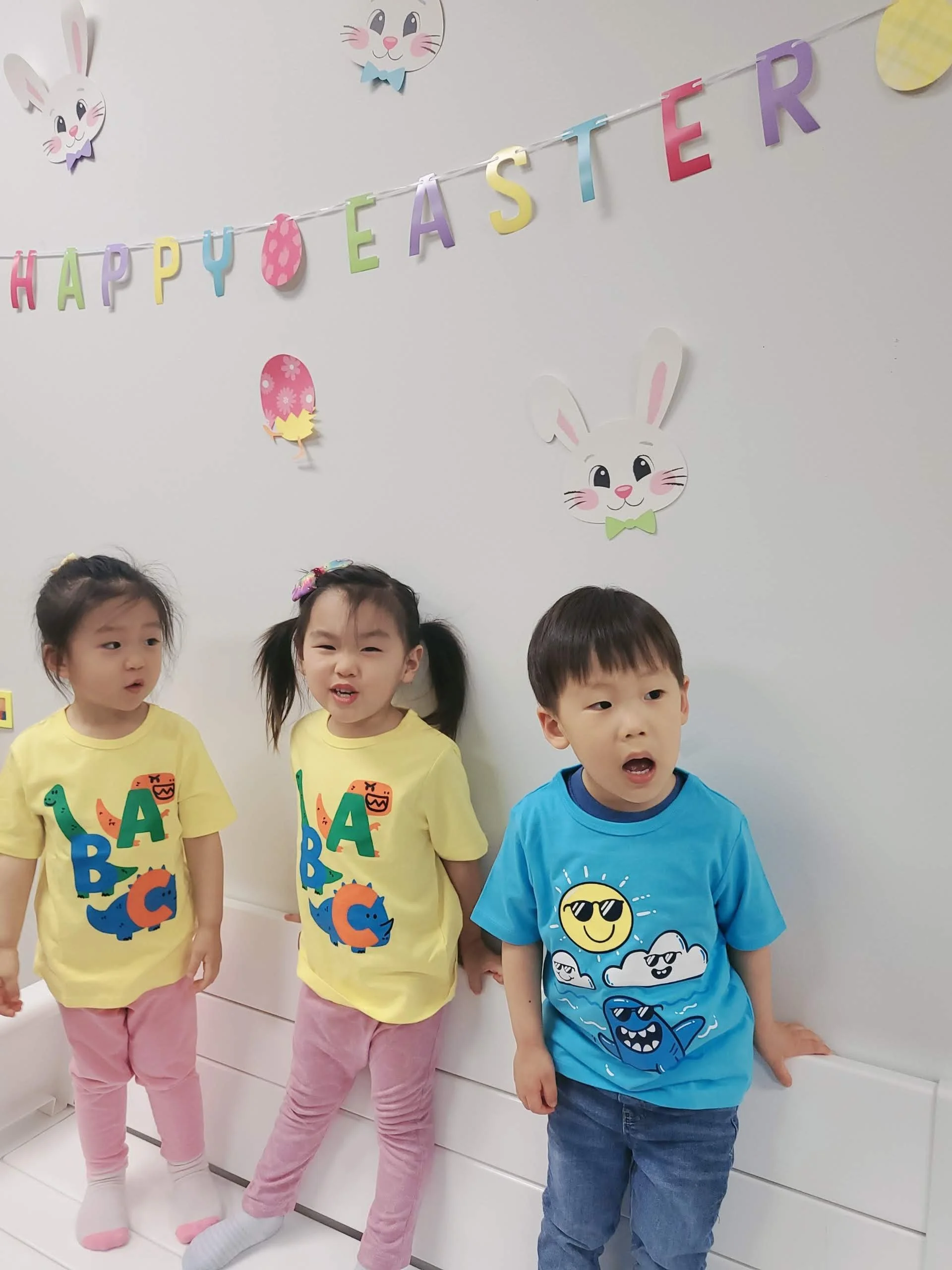 Four children standing in front of a wall decorated with colorful paper cutouts of Easter bunnies and eggs hanging on a string, celebrating Easter.