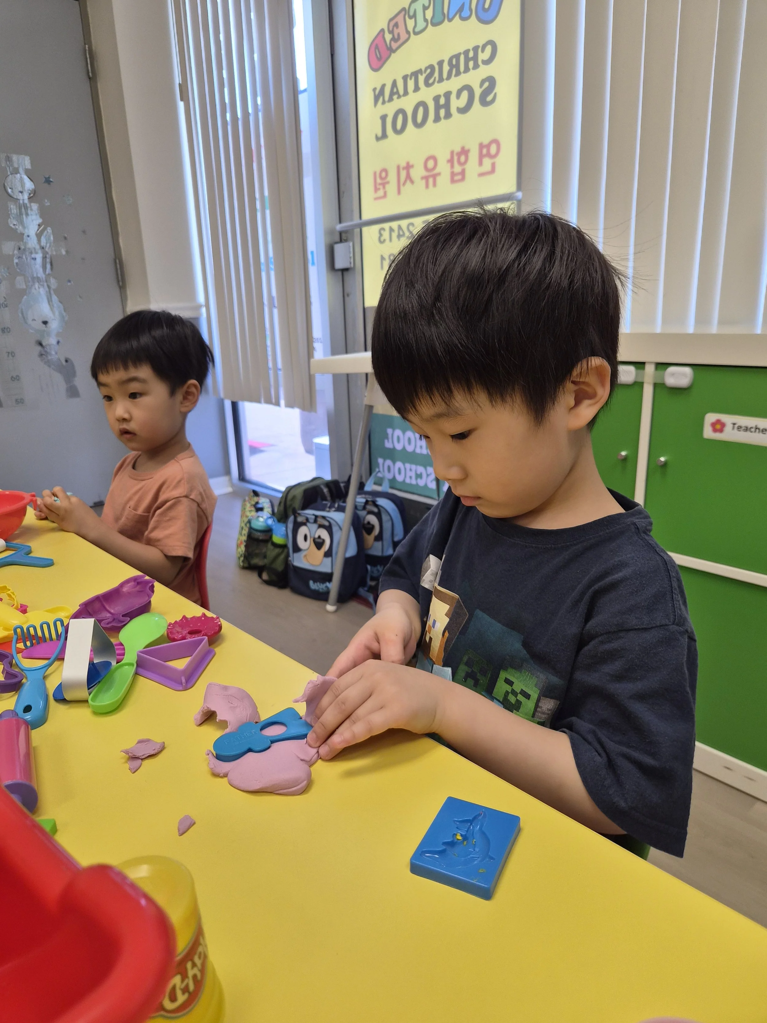 Two young boys are at a yellow table playing with colorful toys and modeling clay. One boy is wearing a pink shirt and is focused on his toys, while the other boy, dressed in a dark shirt, is looking down at his modeling clay.