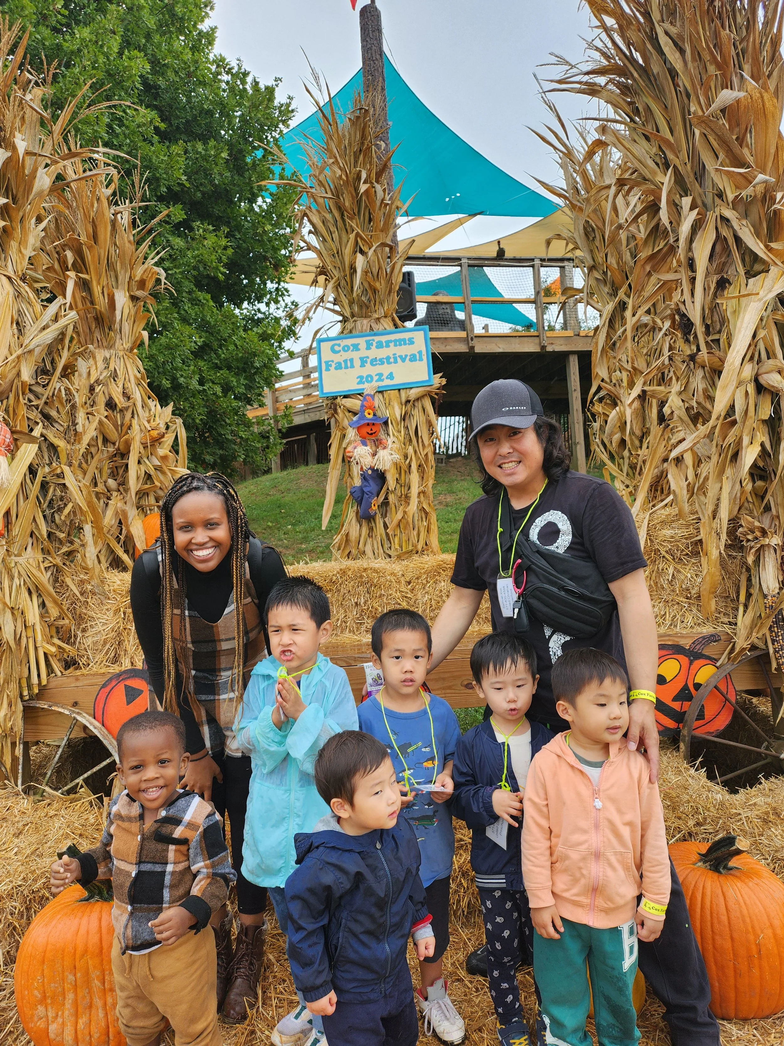 A group of children and two adults posing in front of a pumpkin display at a fall festival. The display includes hay bales, pumpkins, dried cornstalks, and a sign that reads "Cox Farms Fall Festival 2024." There is a wooden platform with a canopy in the background.