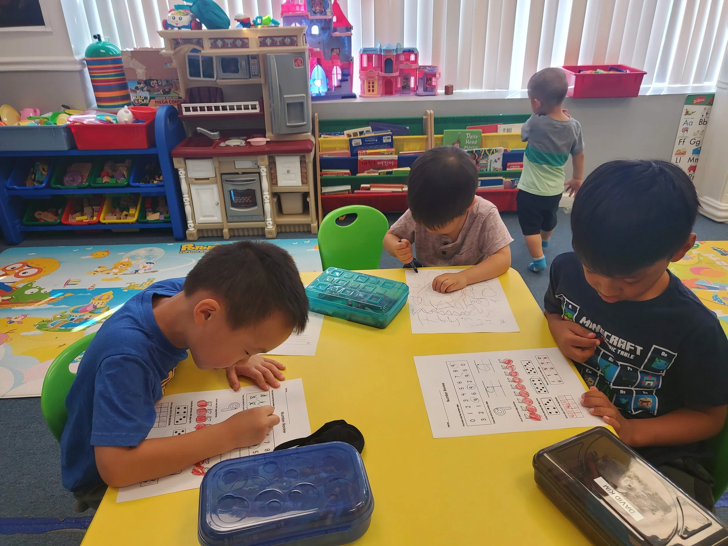Four children sitting at a yellow table doing worksheets, with toys and books on shelves behind them in a playroom.