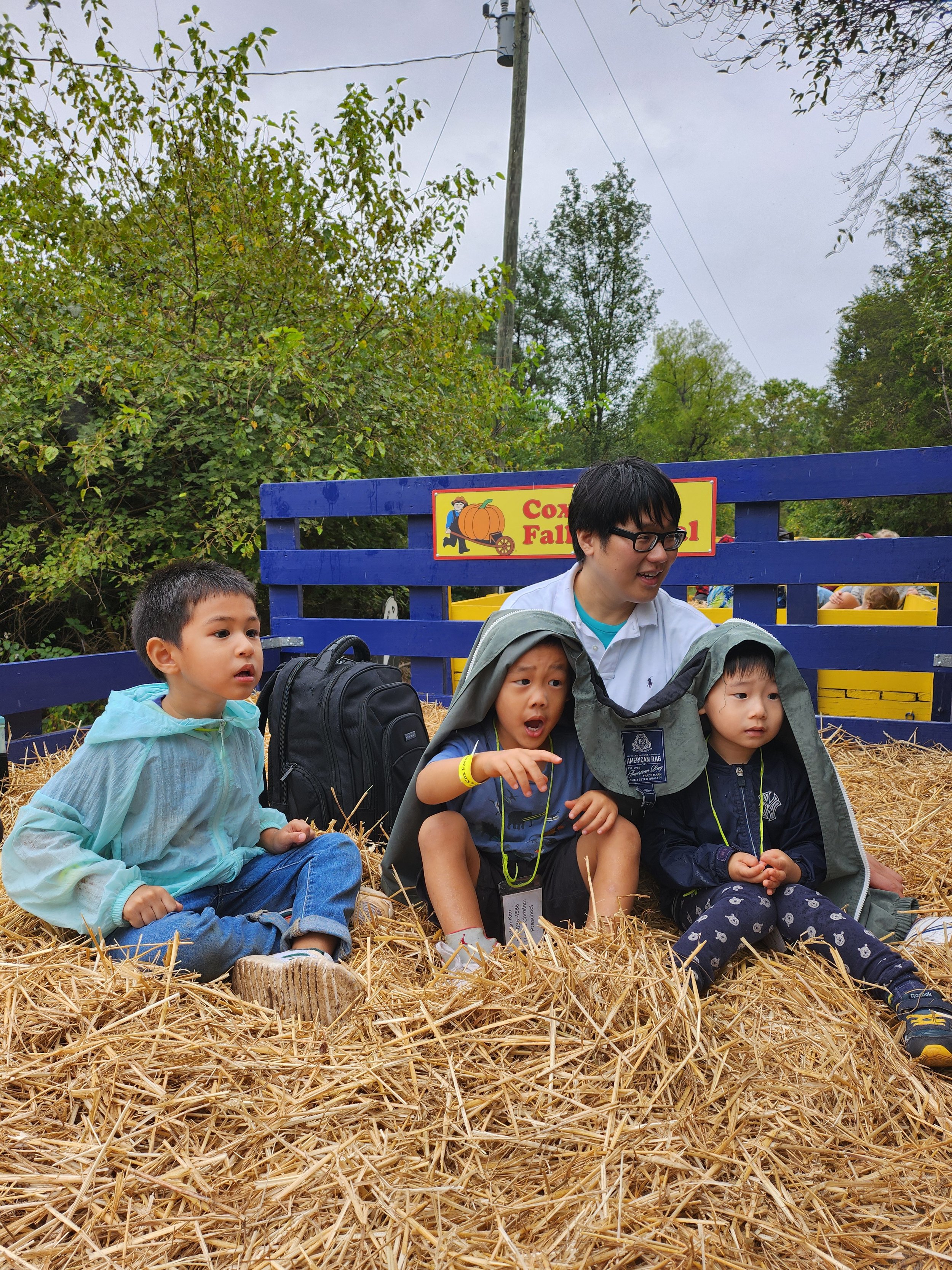 Children and a young man sitting on straw in front of a blue wooden fence at a fall festival or pumpkin patch, with trees and utility poles in the background.