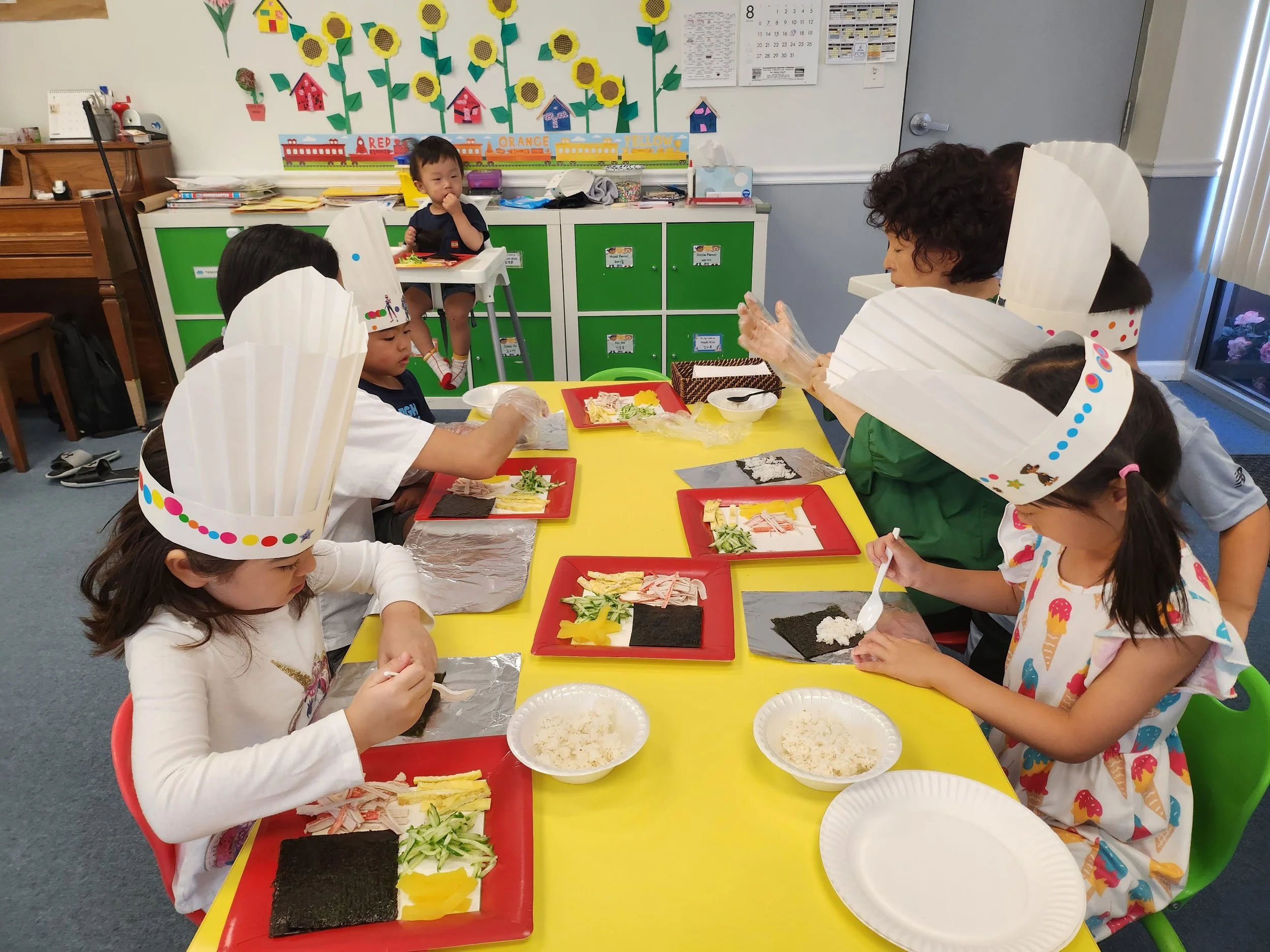 Children and a teacher sitting at a yellow table in a classroom making sushi. The table has red trays with ingredients like rice, seaweed, yellow egg, cucumber, and sliced meat. The children wear chef hats decorated with colorful dots.