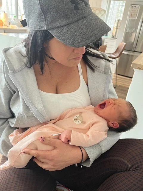 Woman wearing a gray cap and hoodie holding a yawning newborn baby in a pink outfit in a kitchen setting.