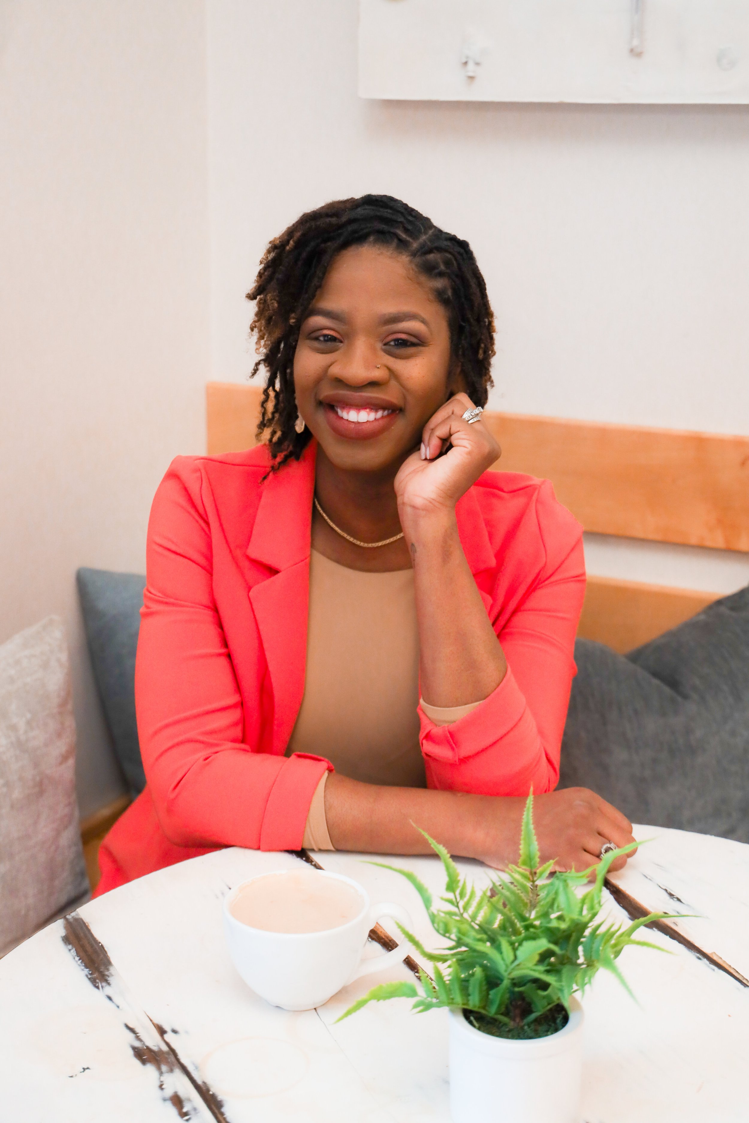 Smiling woman wearing a coral blazer seated at a table with a cup of coffee and a plant.