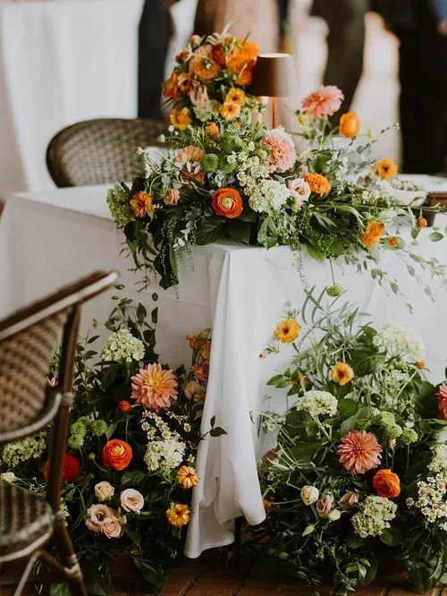 A table decorated with a lush arrangement of colorful flowers in shades of orange, pink, white, and green, with a small brown lampshade on top.