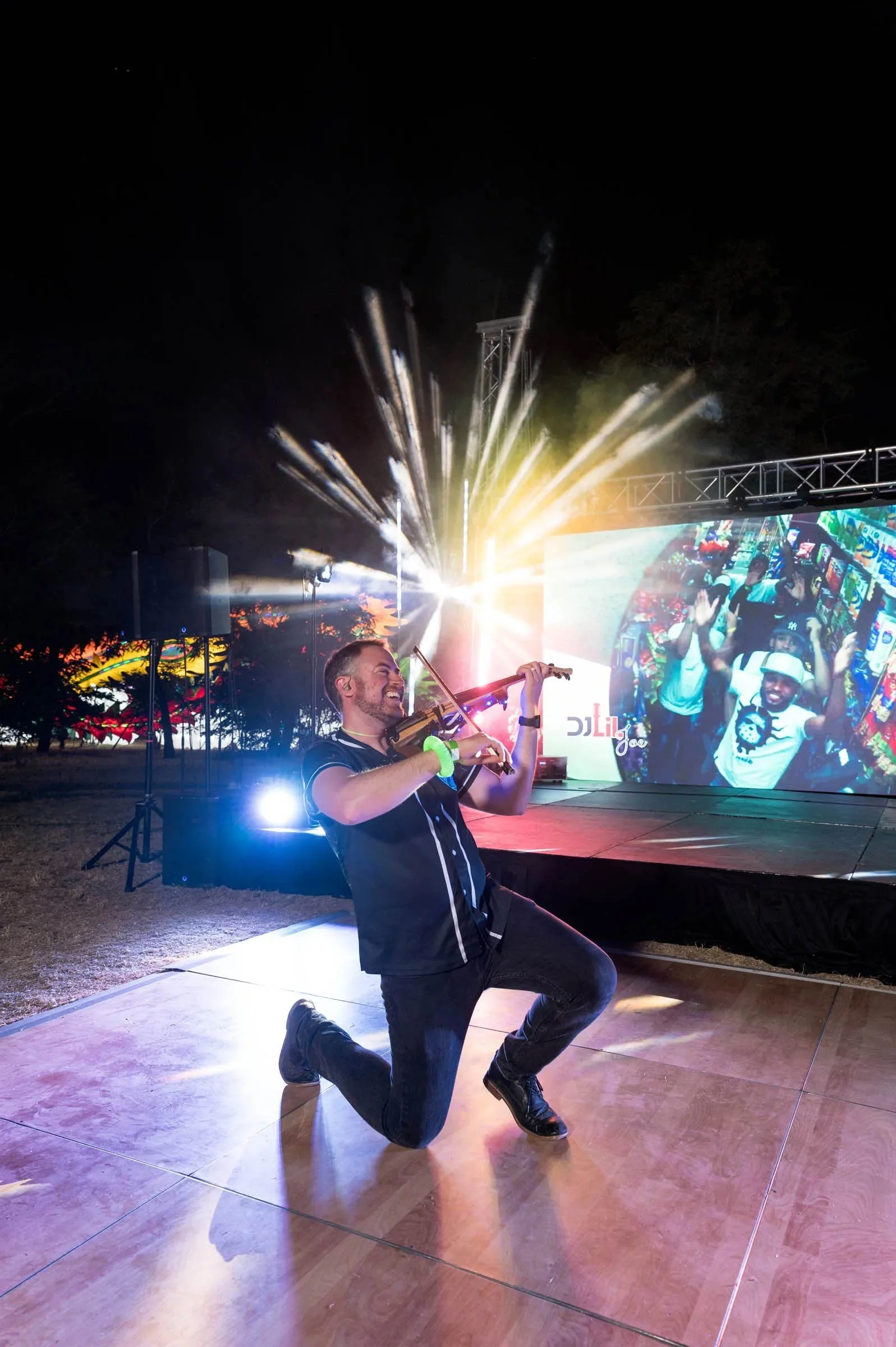 A man kneeling and playing a violin on a dance floor at night with colorful stage lights and a large screen displaying a crowd in the background.