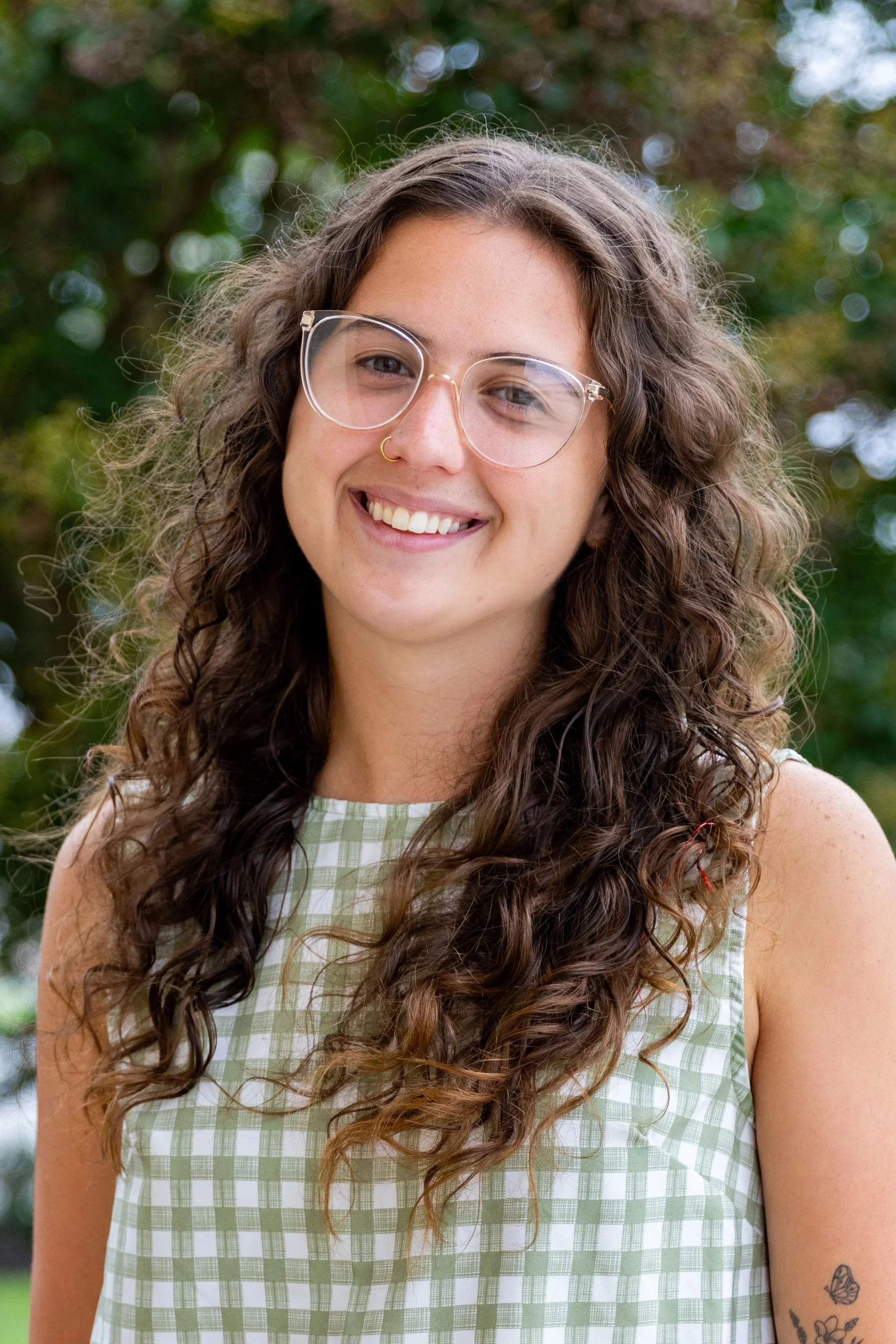 A young woman with curly brown hair, glasses, and a nose ring smiling outdoors.