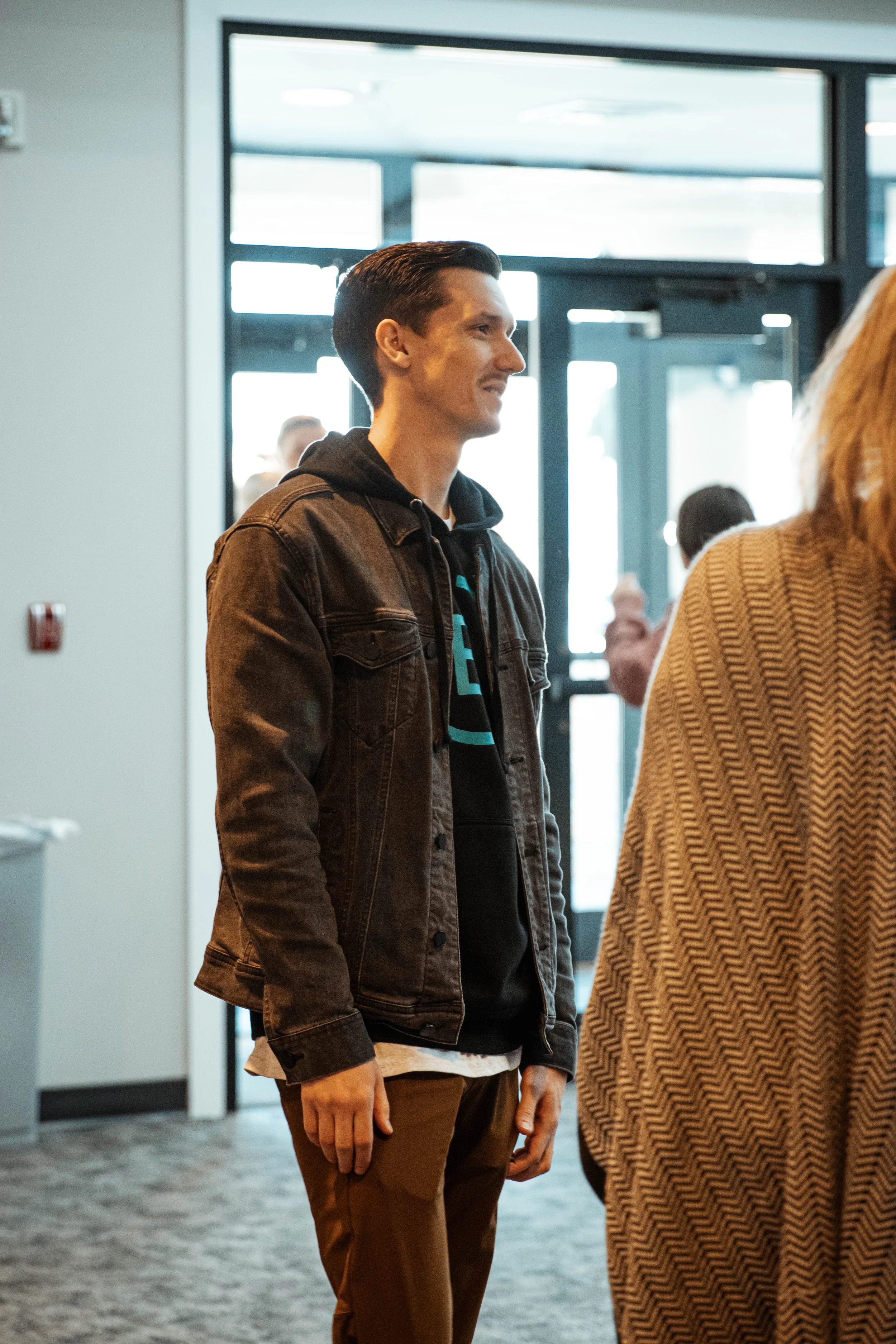 A young man standing indoors, wearing a brown denim jacket over a black hoodie and brown pants, smiling and looking to his left.
