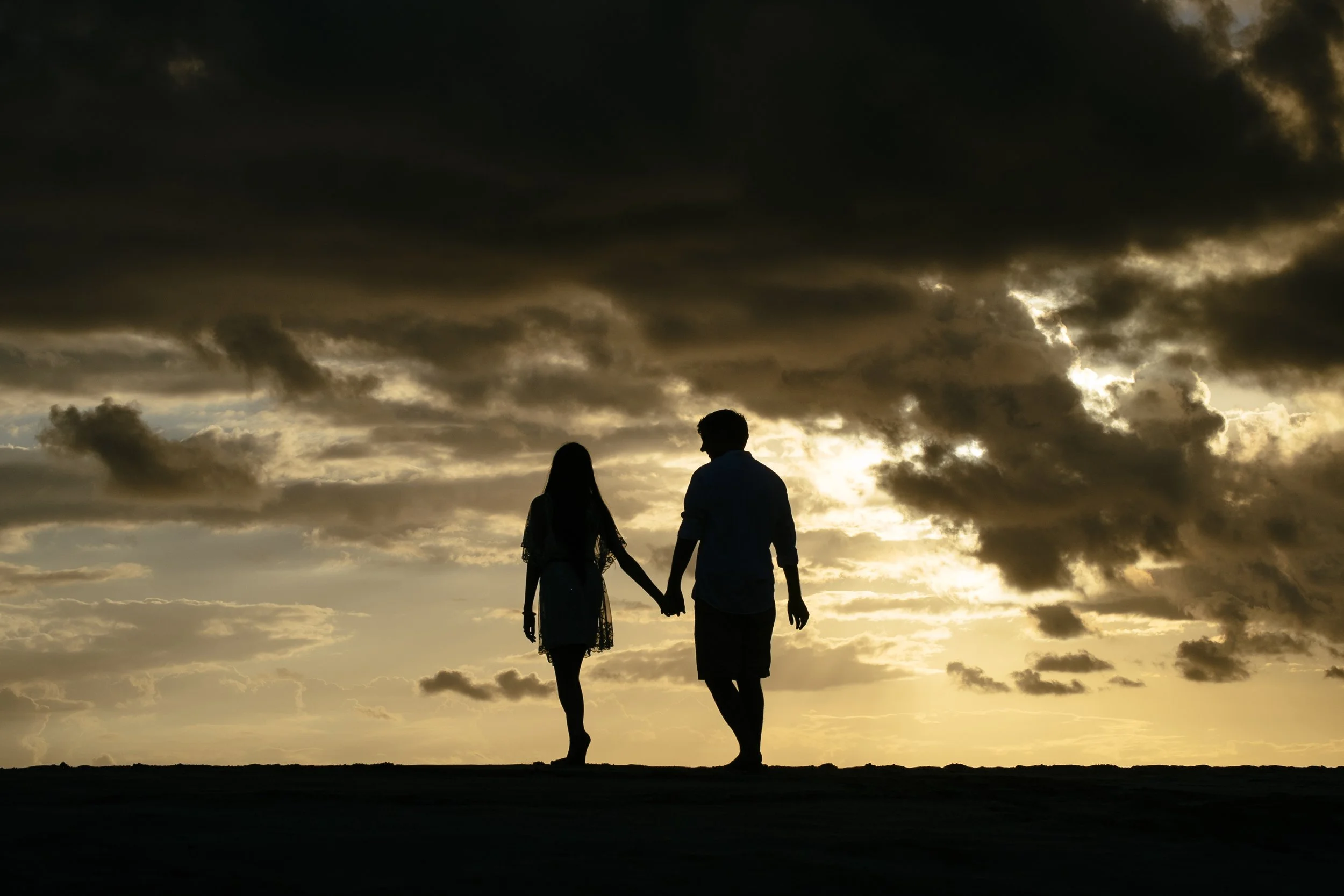 Silhouetted couple holding hands as they walk toward a glowing ocean sunset.