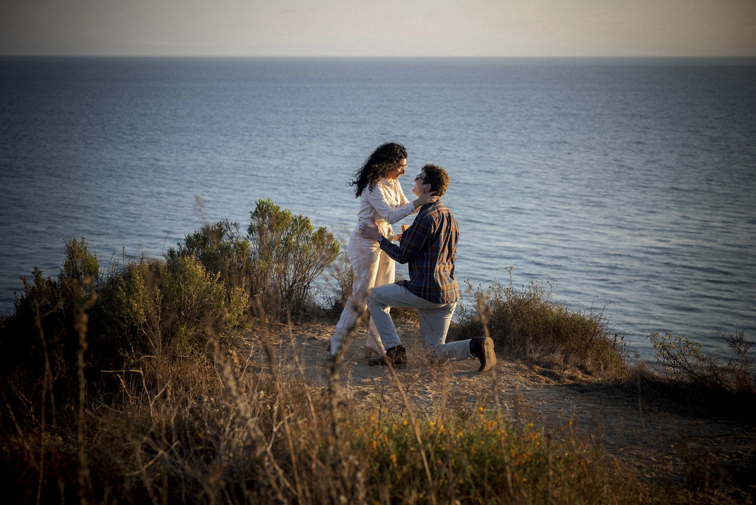 Man kneeling during a surprise proposal on coastal bluffs above the ocean.