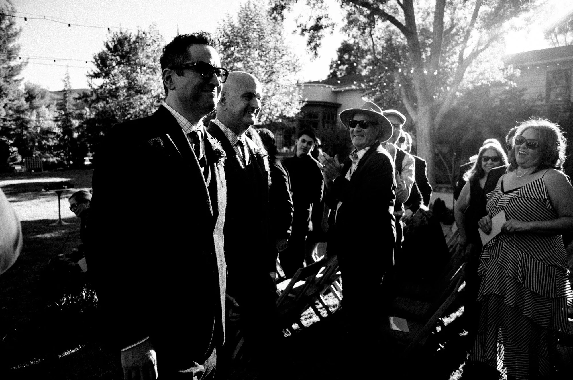 Two grooms standing together before guests during an outdoor wedding ceremony.