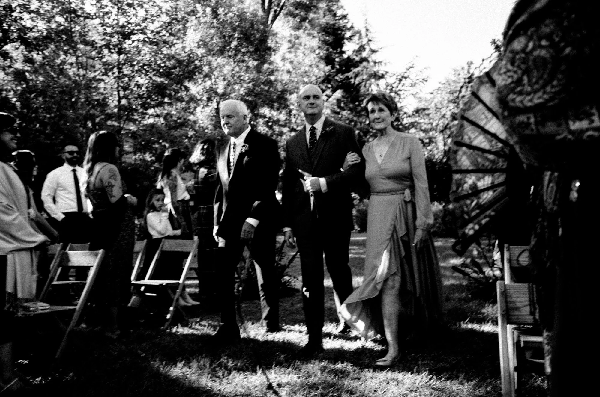 Groom and parents walking down aisle during an outdoor wedding ceremony.