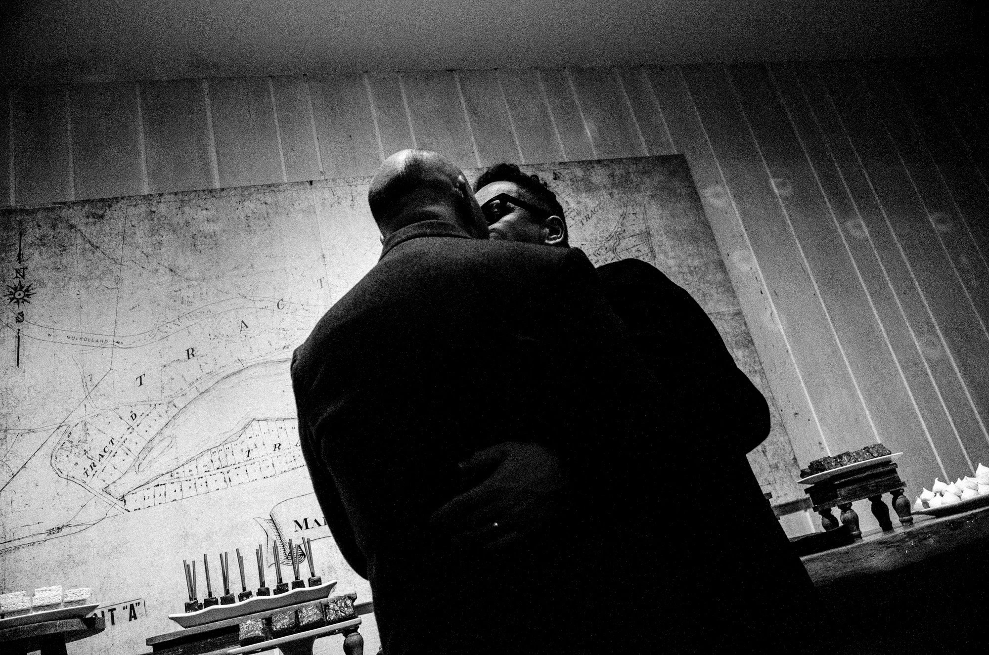 Two grooms embracing beneath a large wall map in black and white.