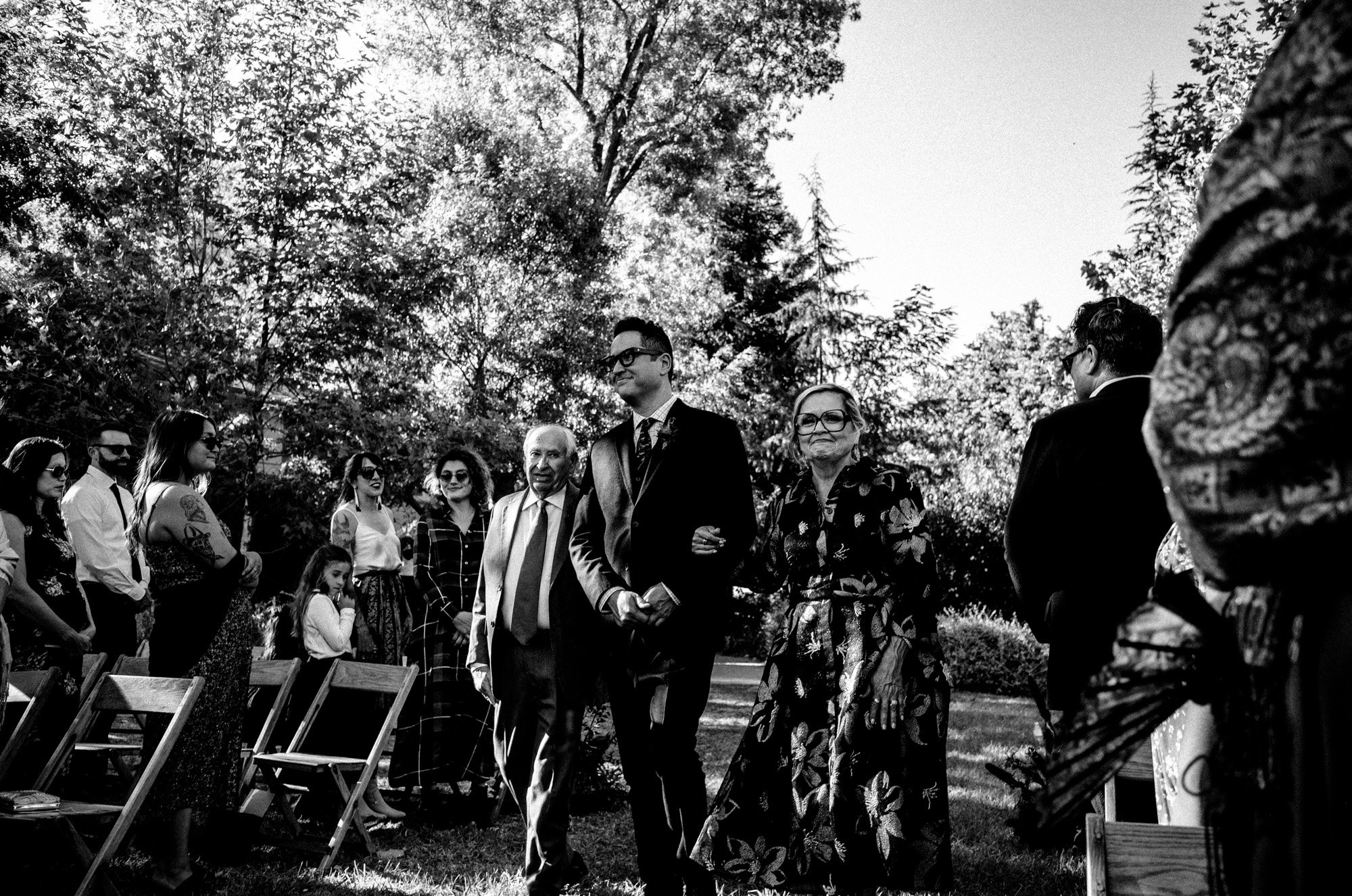 Groom and parents walking down aisle during an outdoor wedding ceremony.