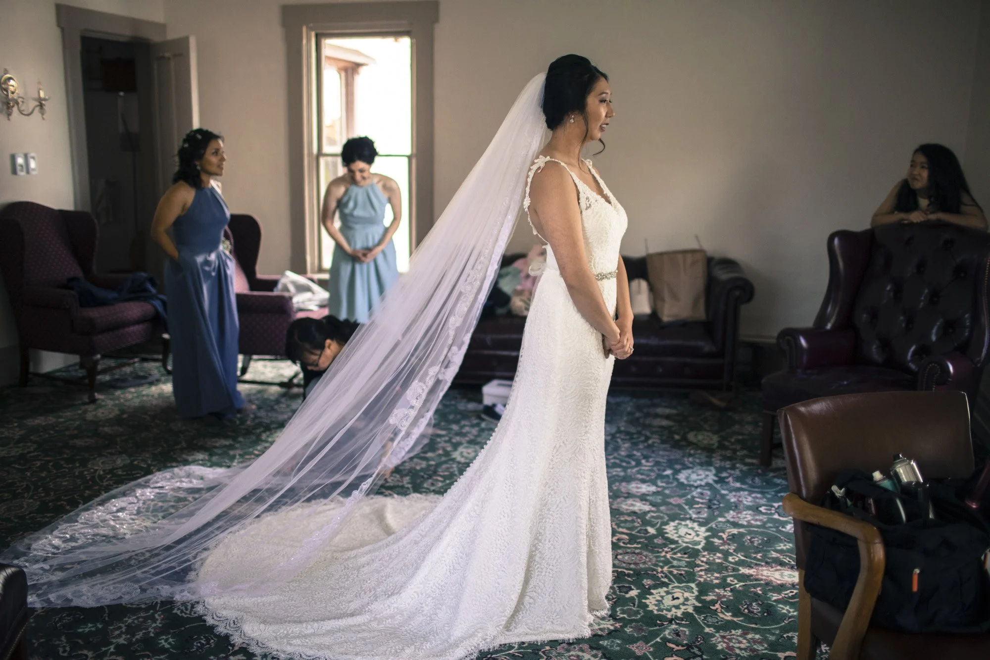 Bride standing in profile with her long veil as attendants look on.