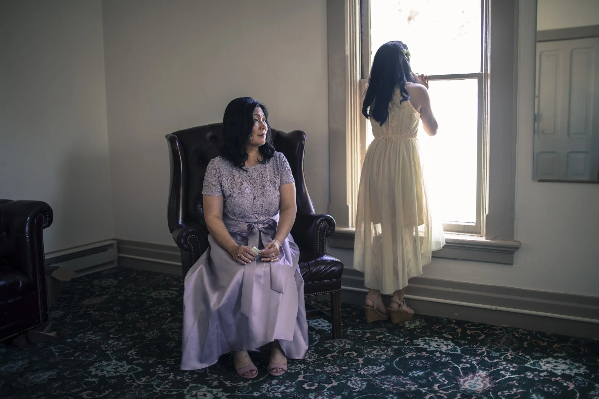 Bride standing near a window while a seated bridesmaid watches.