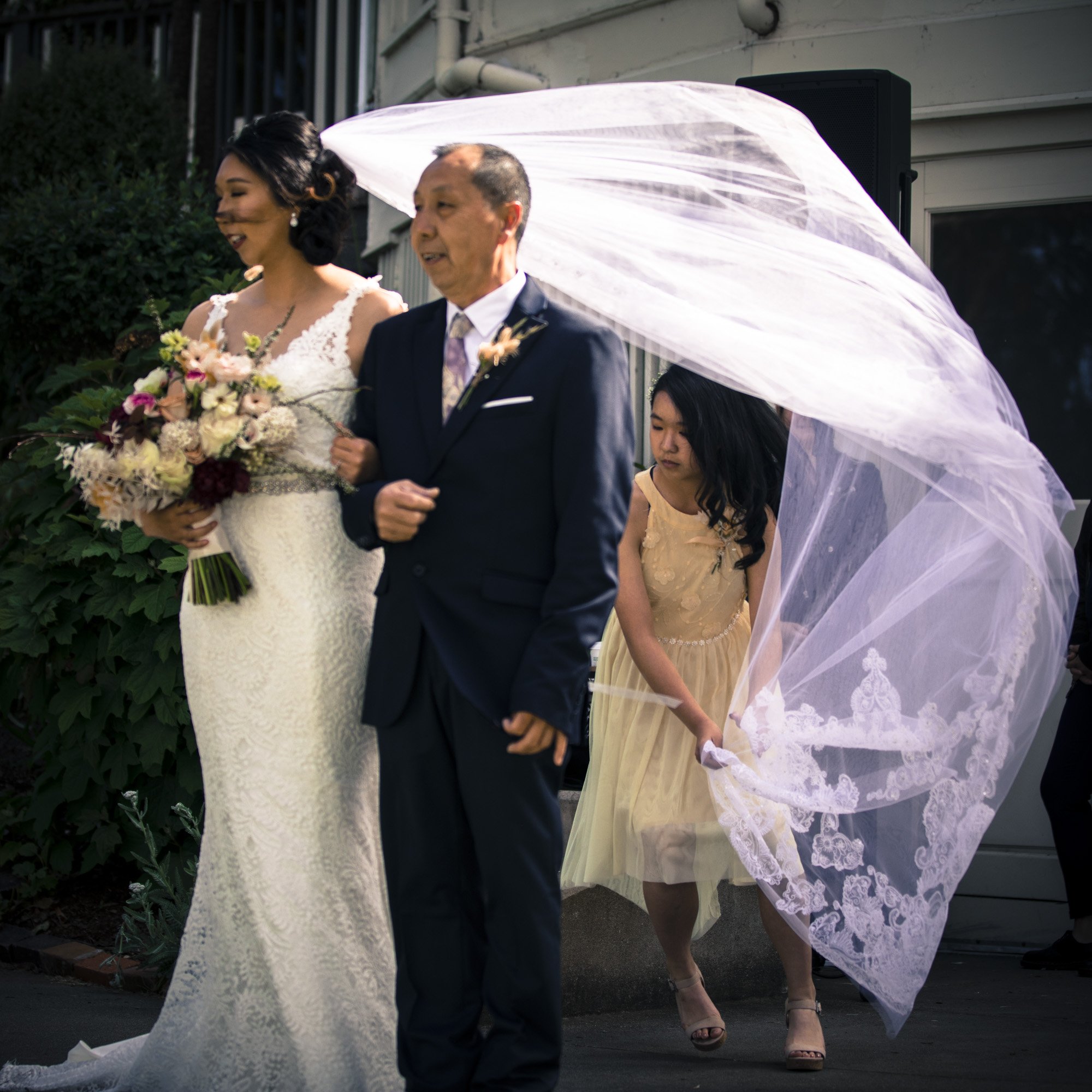 Bride walking outdoors with her father as wind lifts her veil.