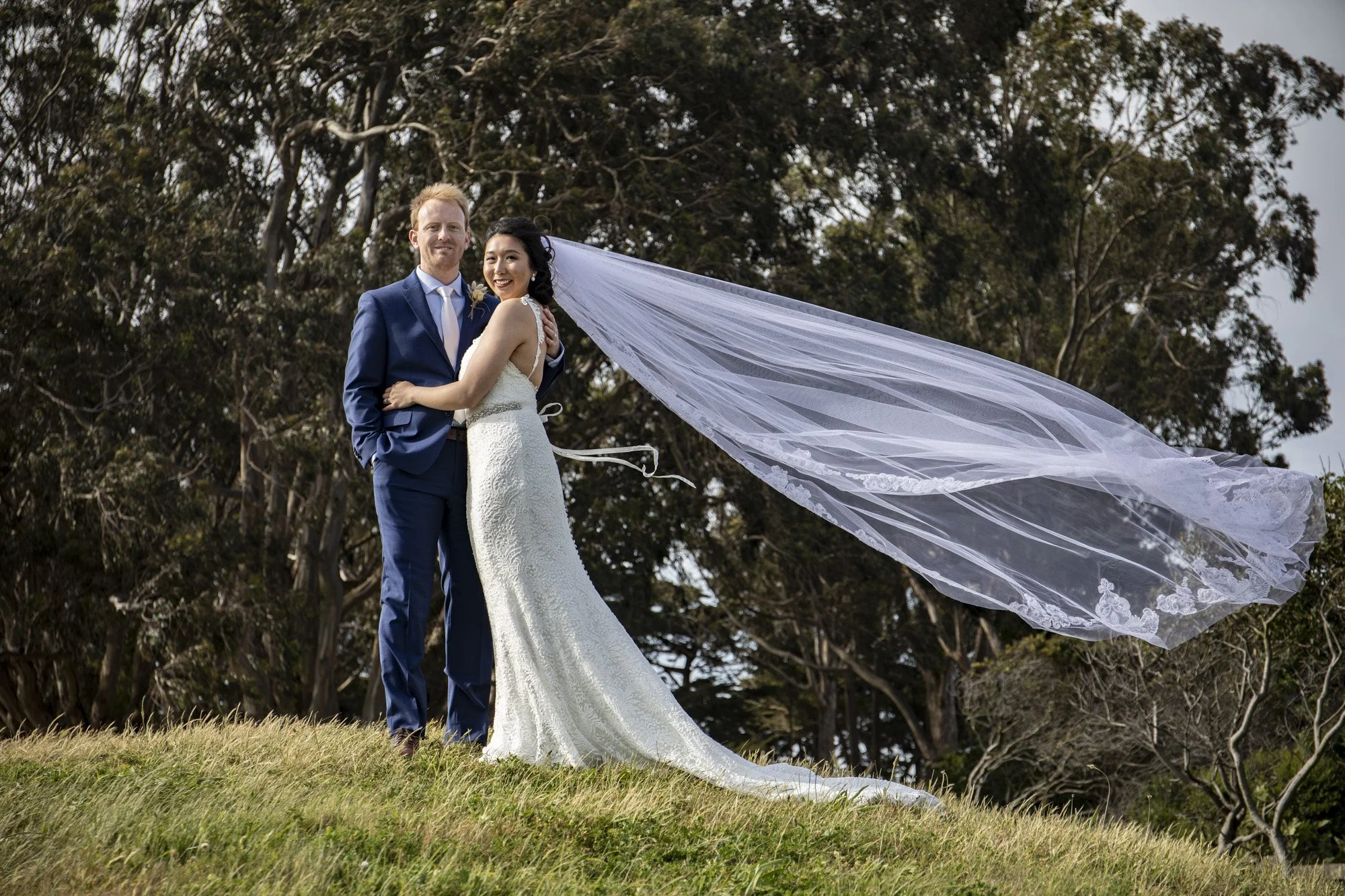 Newlyweds standing together as the bride’s veil streams in the wind.