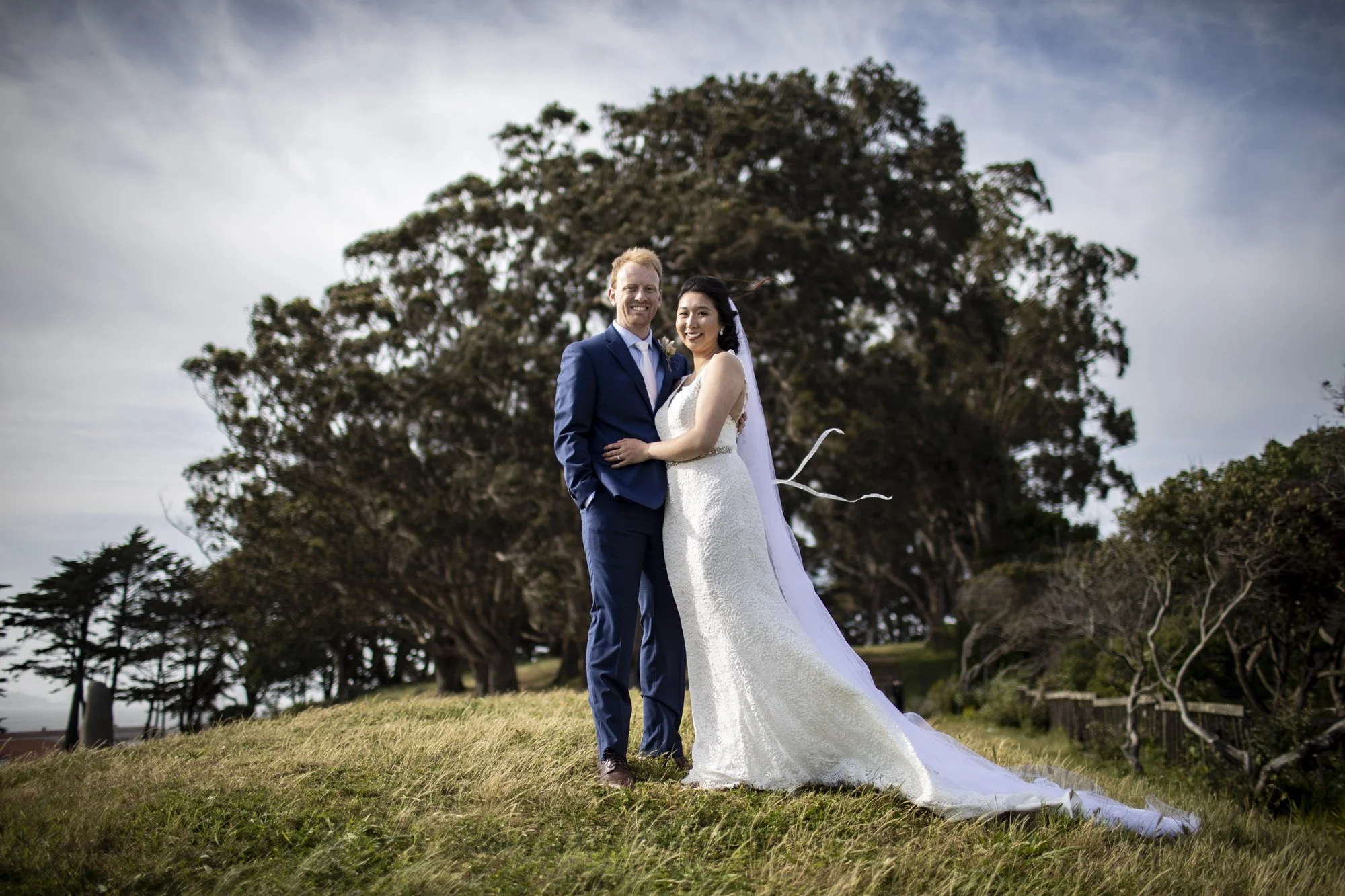 Newlyweds standing close together beneath a broad oak tree.