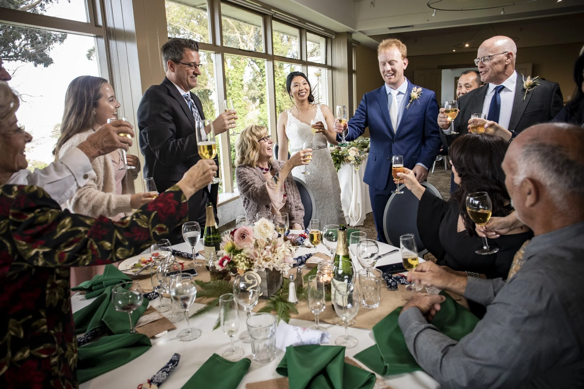 Newlyweds and guests raising glasses around a reception table.