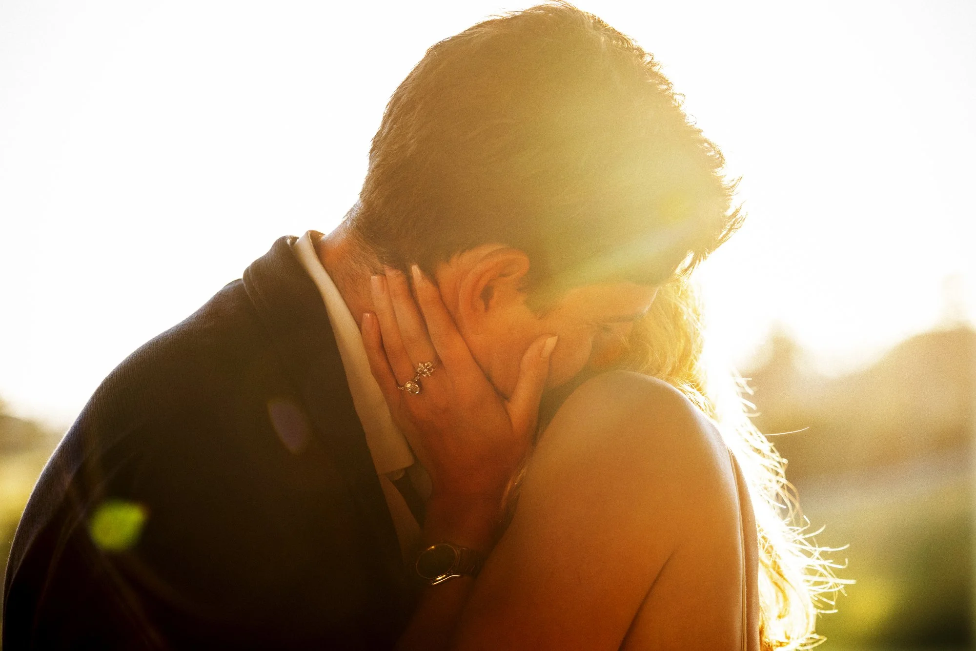Close-up of a newly engaged couple kissing in golden backlight, with the engagement ring visible.