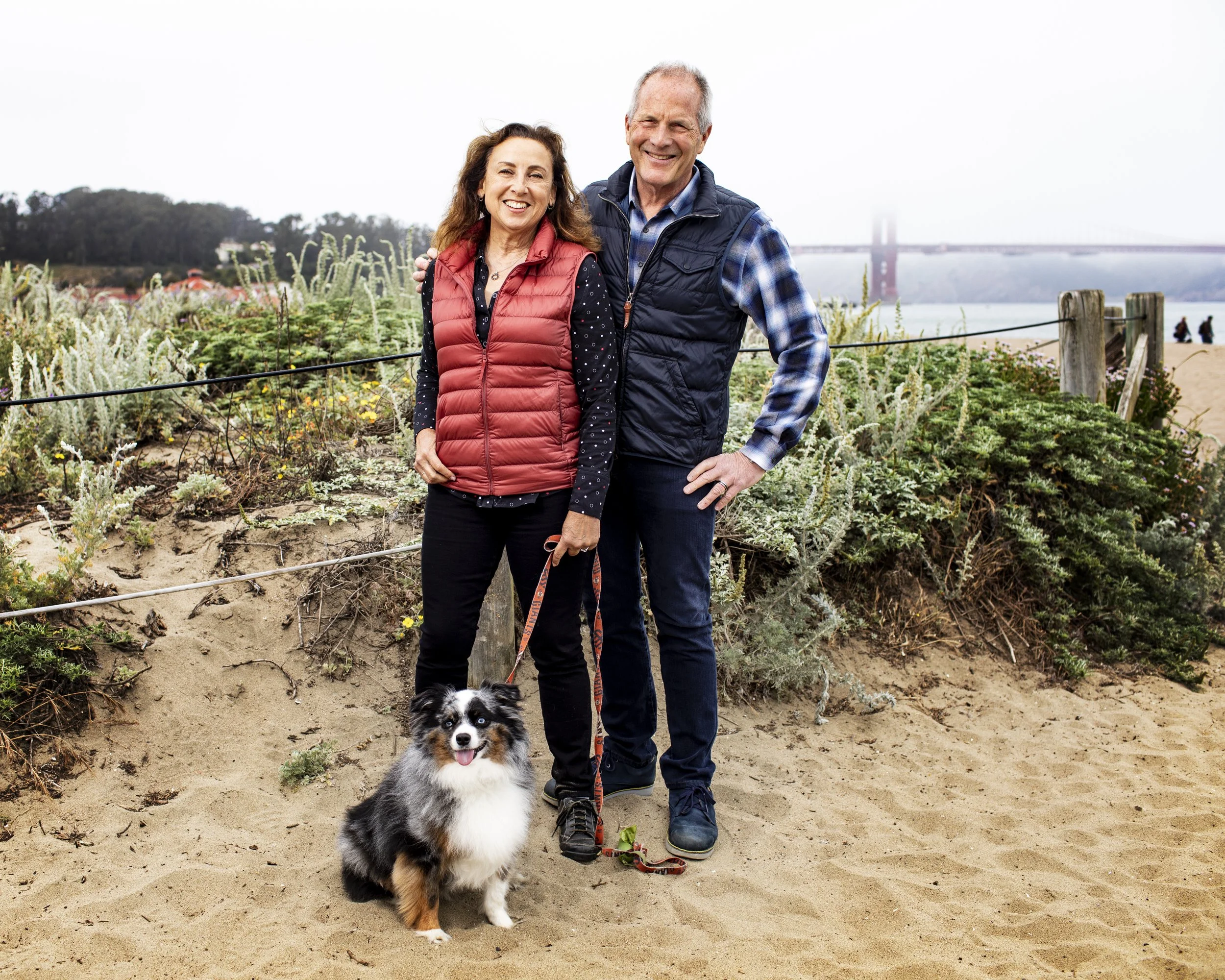 Couple standing with their dog on a beach near the Golden Gate Bridge.