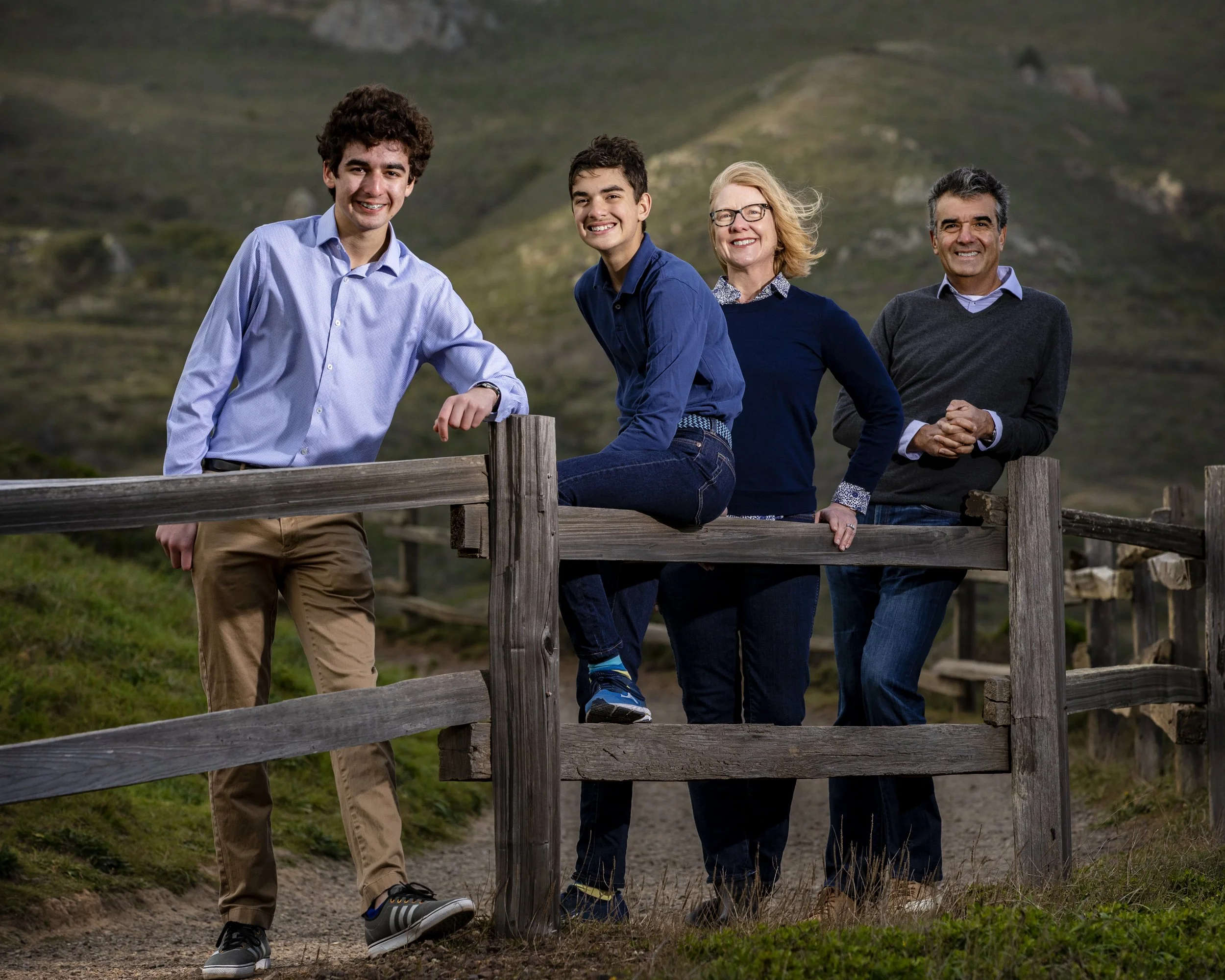Family of four standing together beside a wooden fence in a rural landscape.