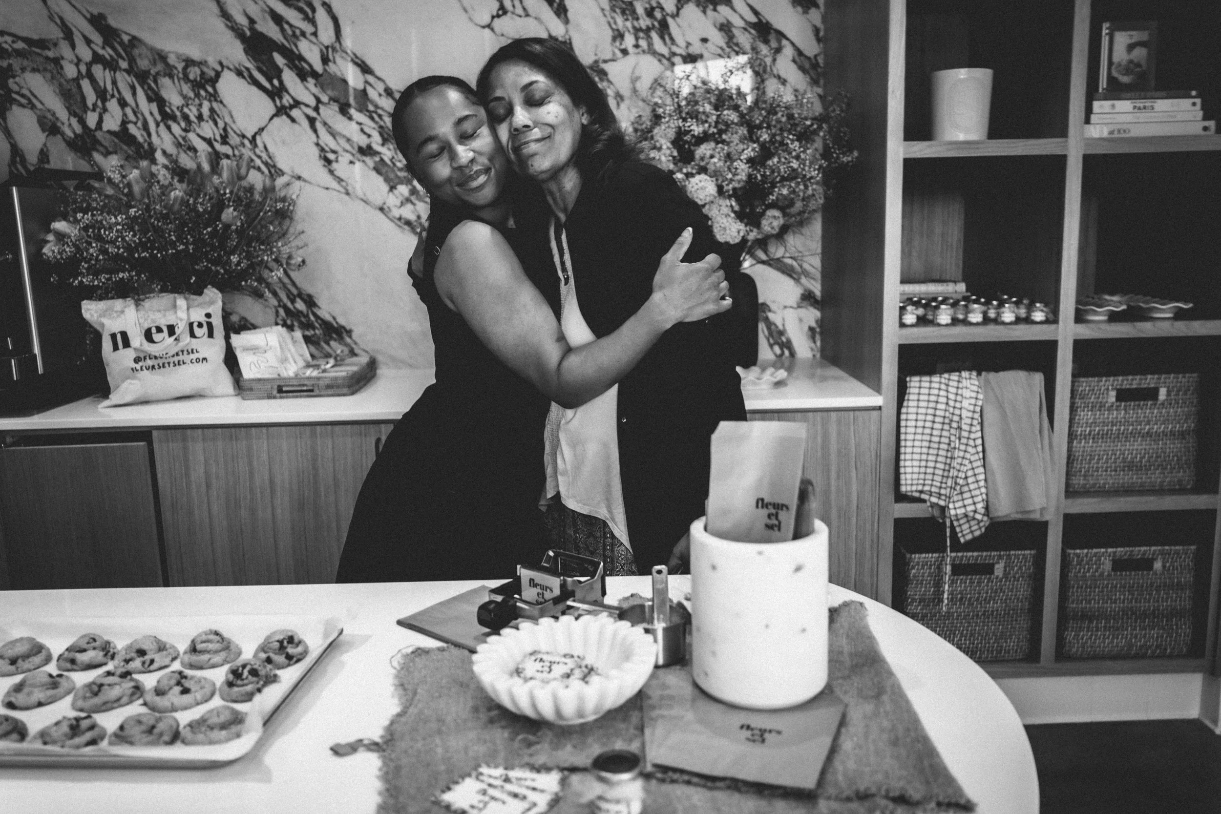 Two women embracing beside a celebration cookie bakery in black and white.