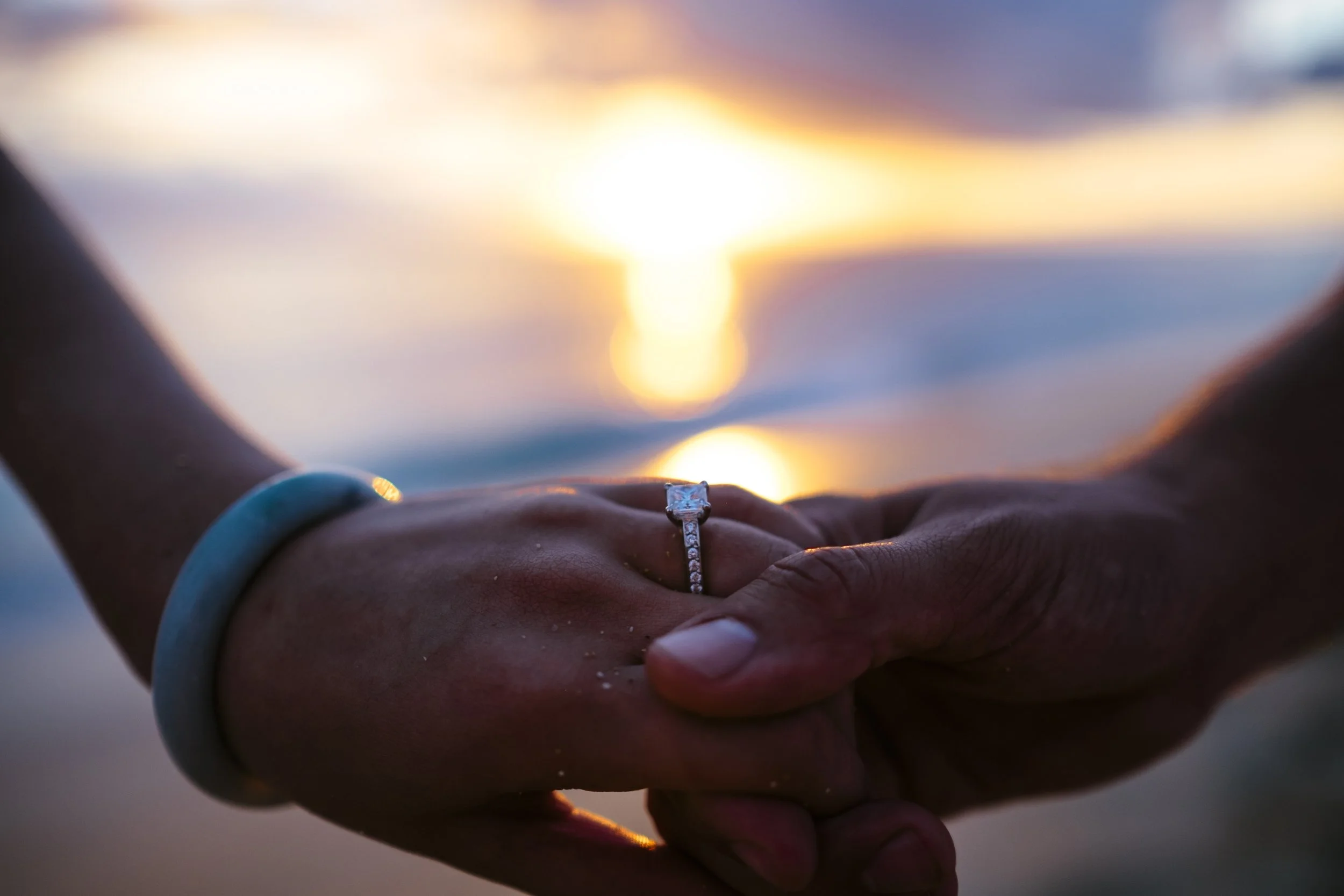 Engagement ring held between a couple’s hands against a blurred sunset.
