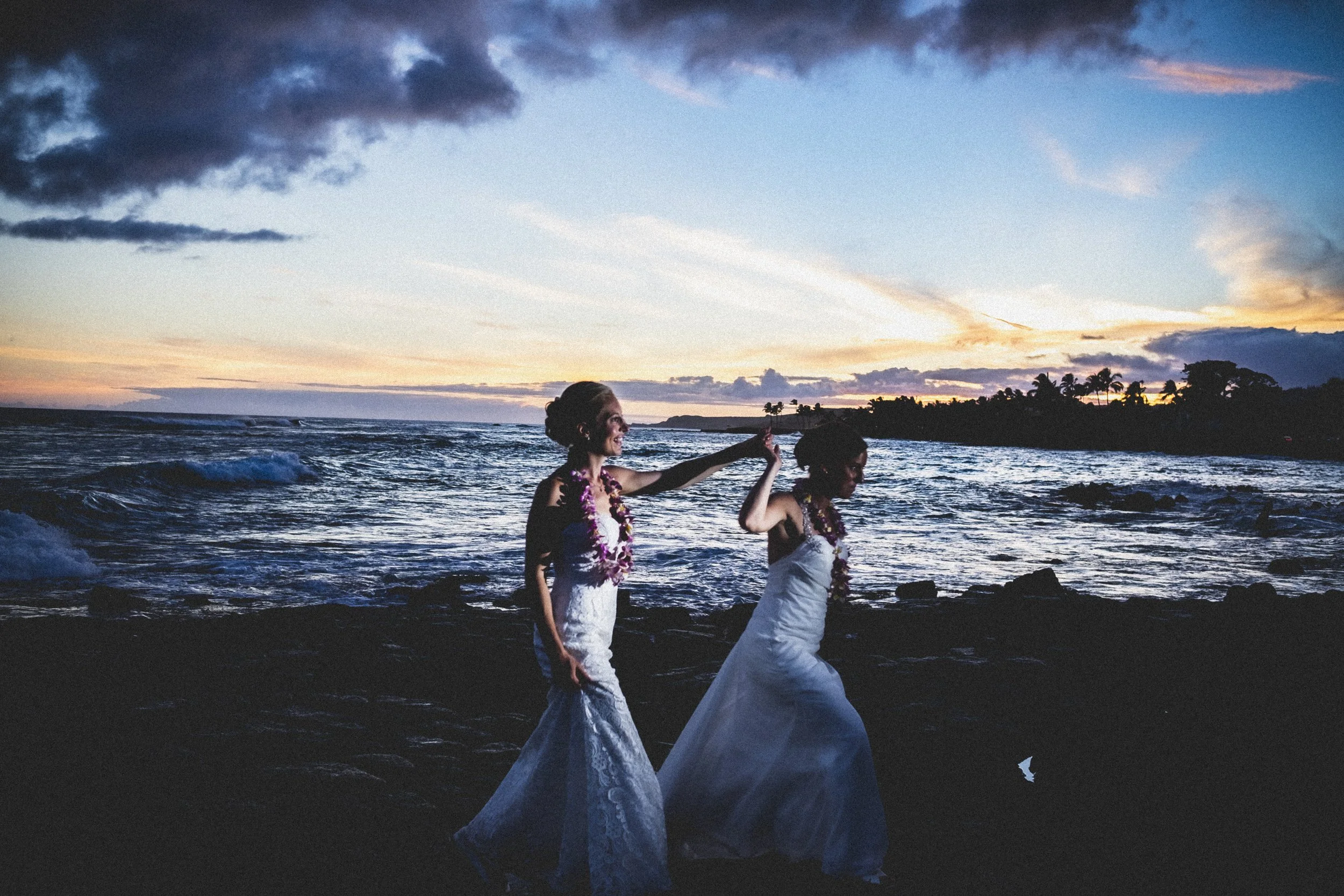 Two brides walking with each other on a rocky shoreline after their wedding.