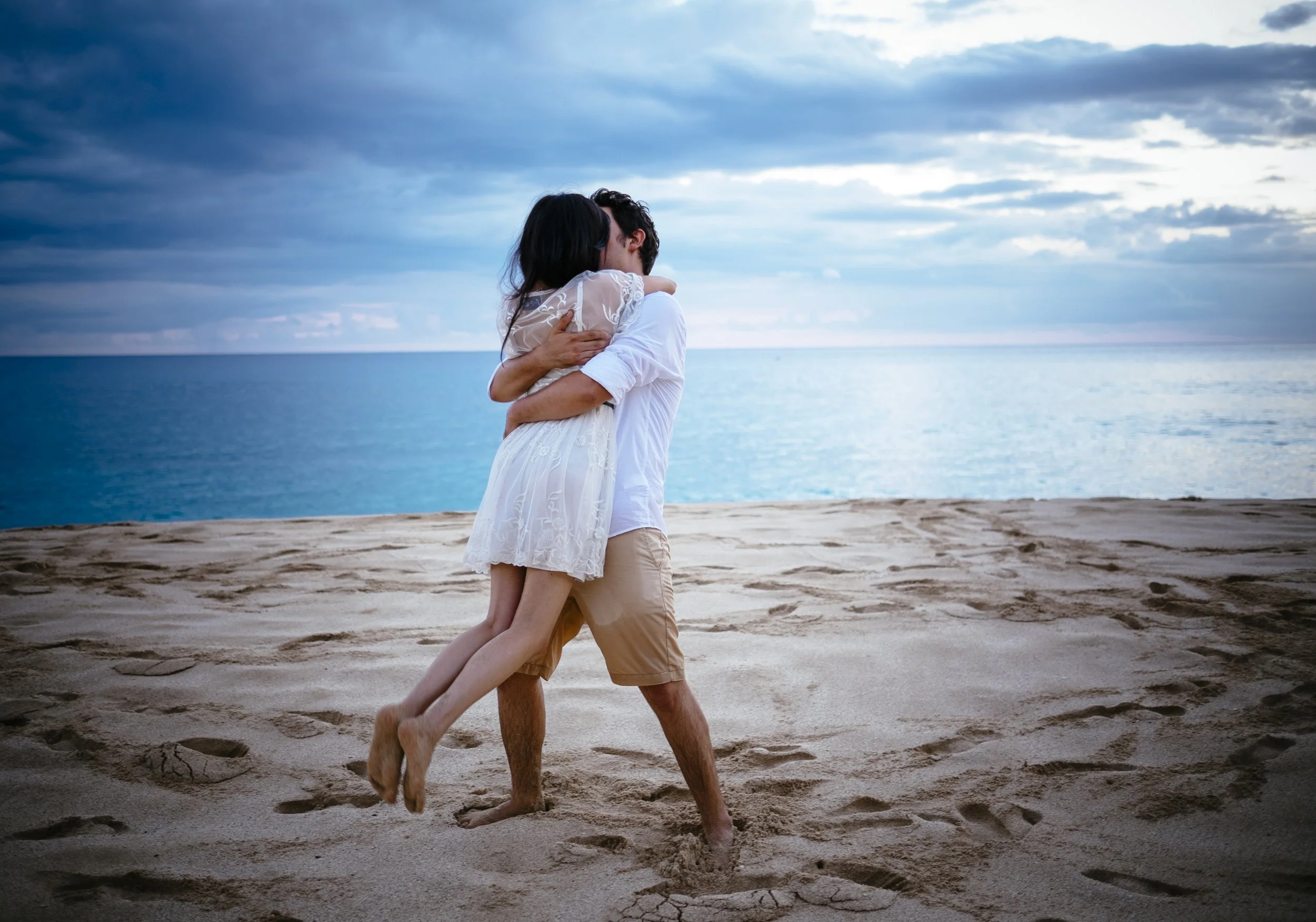 Couple lifting into an embrace on an Oahu beach beneath a dramatic sky.