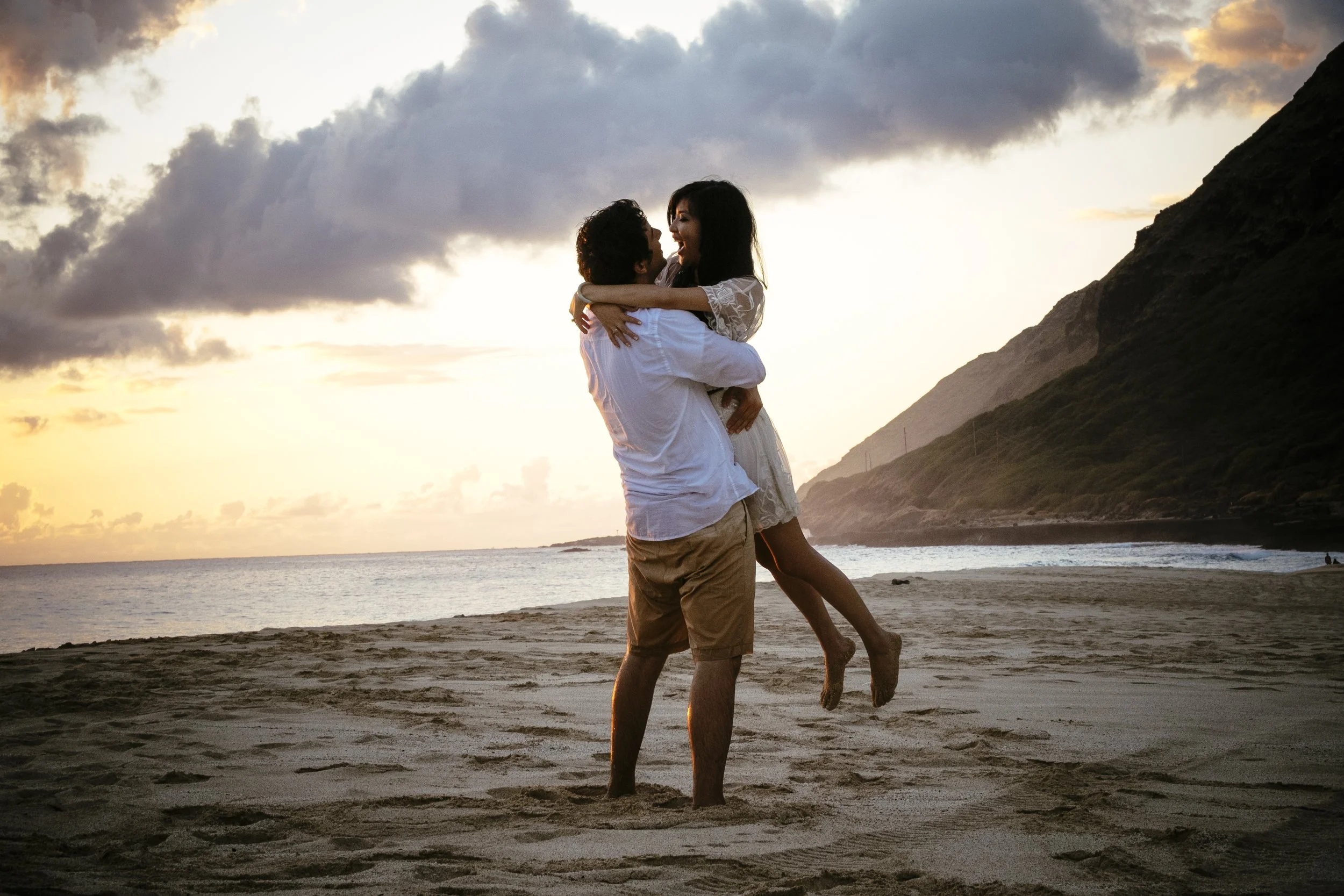 Couple lifting into an embrace on an Oahu beach beneath dramatic mountains.