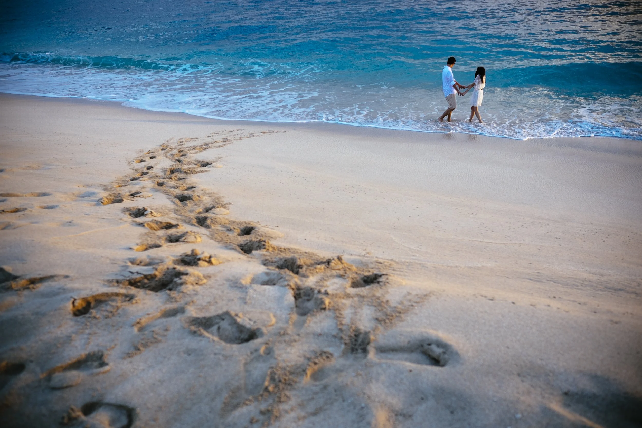 Couple walking hand in hand along an Oahu beach, leaving footprints in the sand.