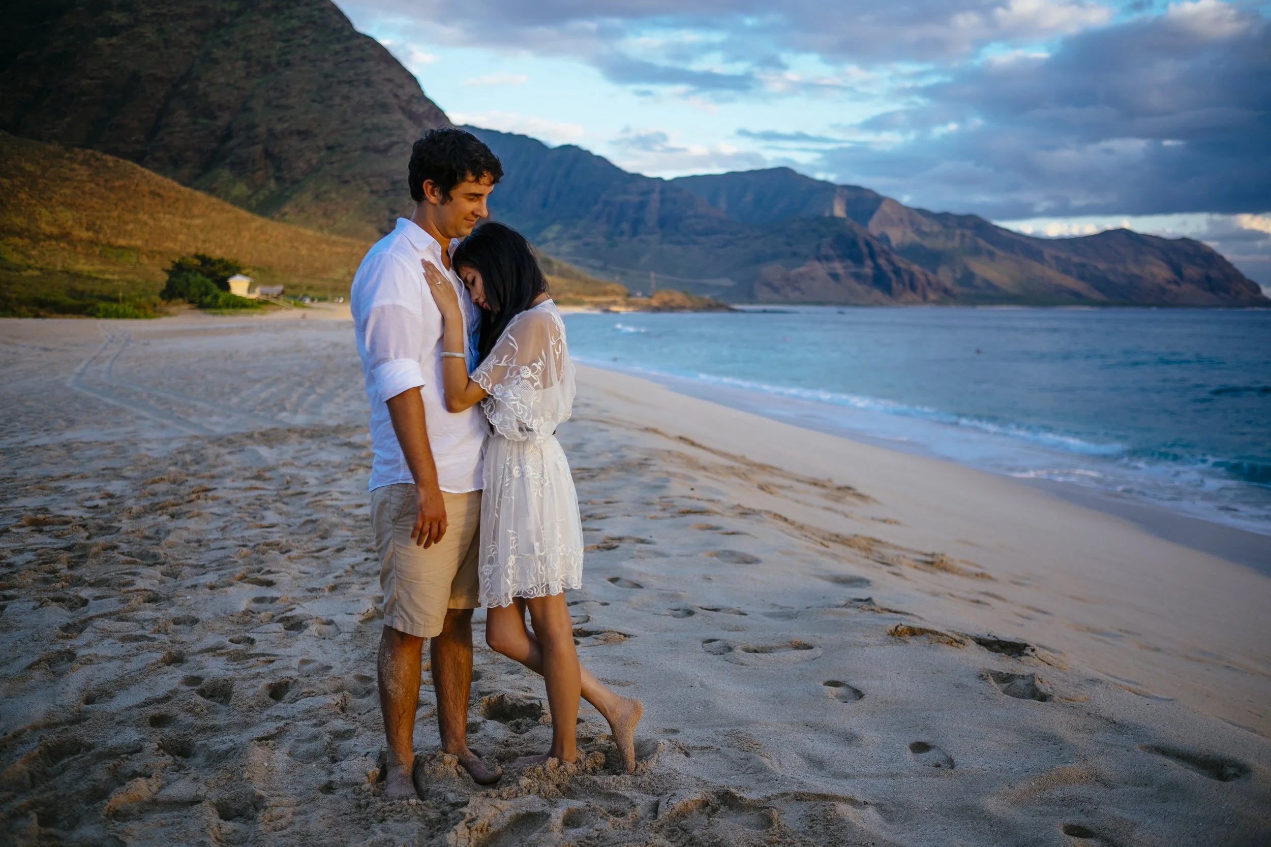 Couple standing together on an Oahu beach with green mountains behind them.
