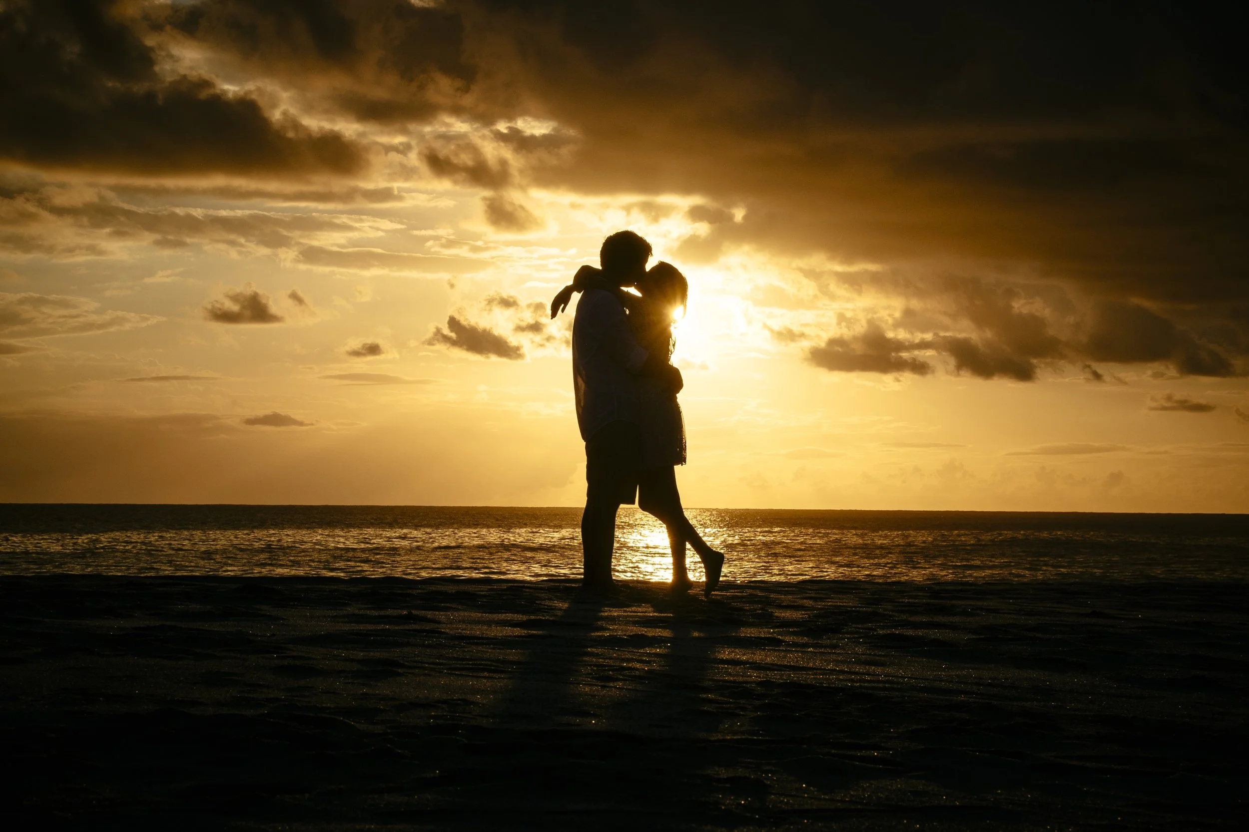 Silhouetted couple embracing at the water’s edge beneath a dramatic sunset.