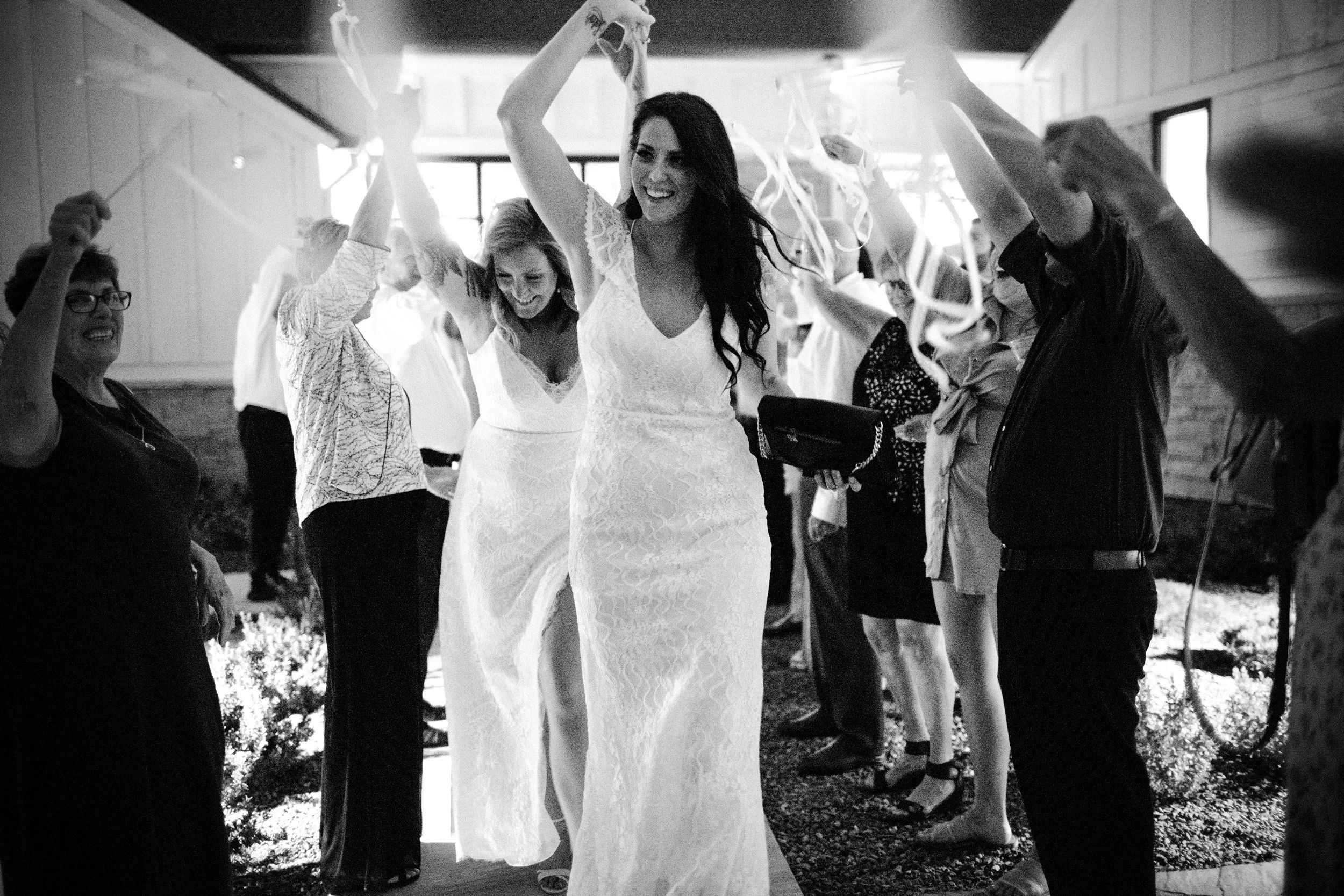 Brides raising their arms among cheering guests during a wedding celebration.
