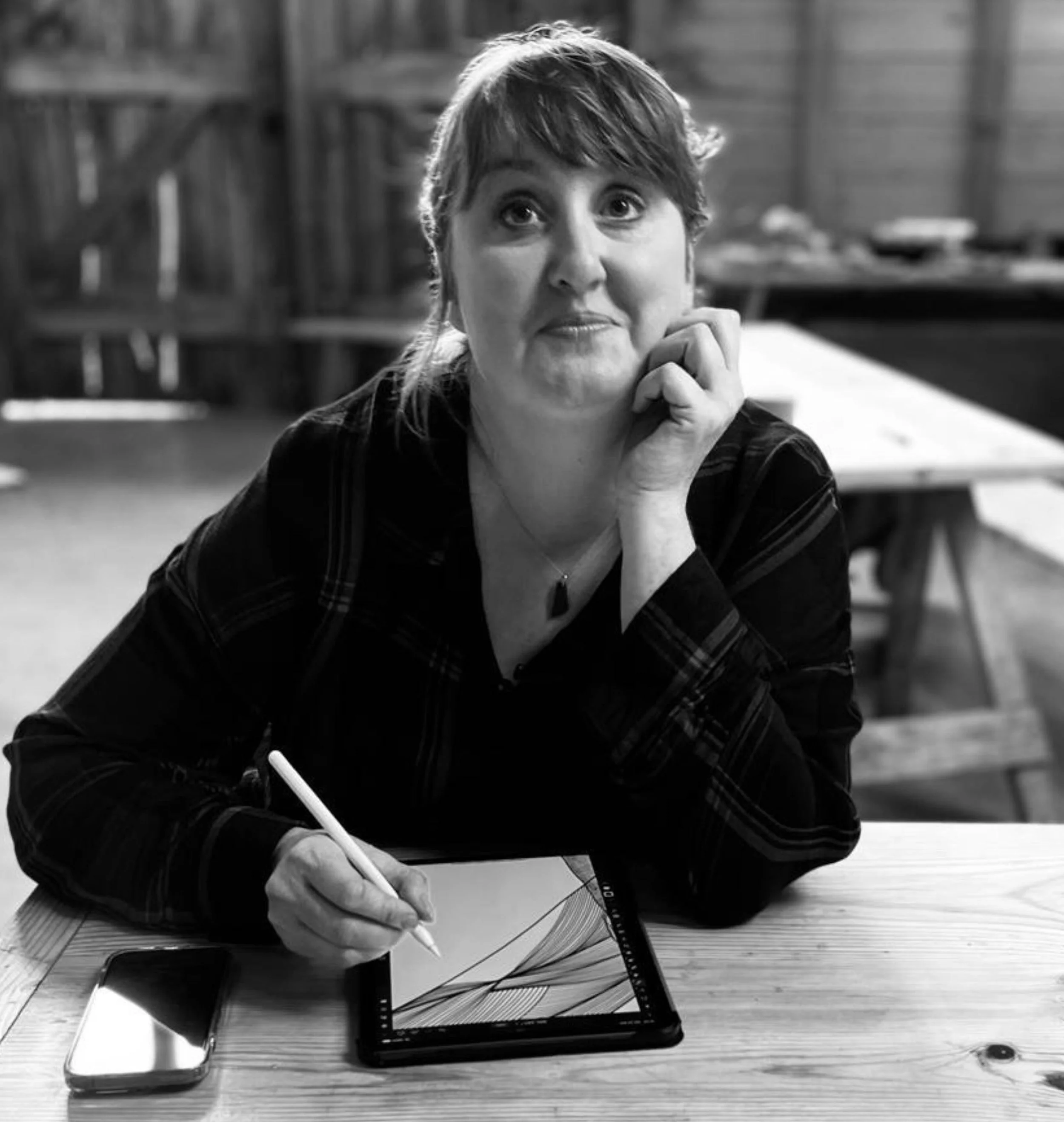 Black and white photo of a woman with short hair, sitting at a table, holding a pen and a notebook, with a laptop and a smartphone on the table, in a rustic setting with wooden walls.