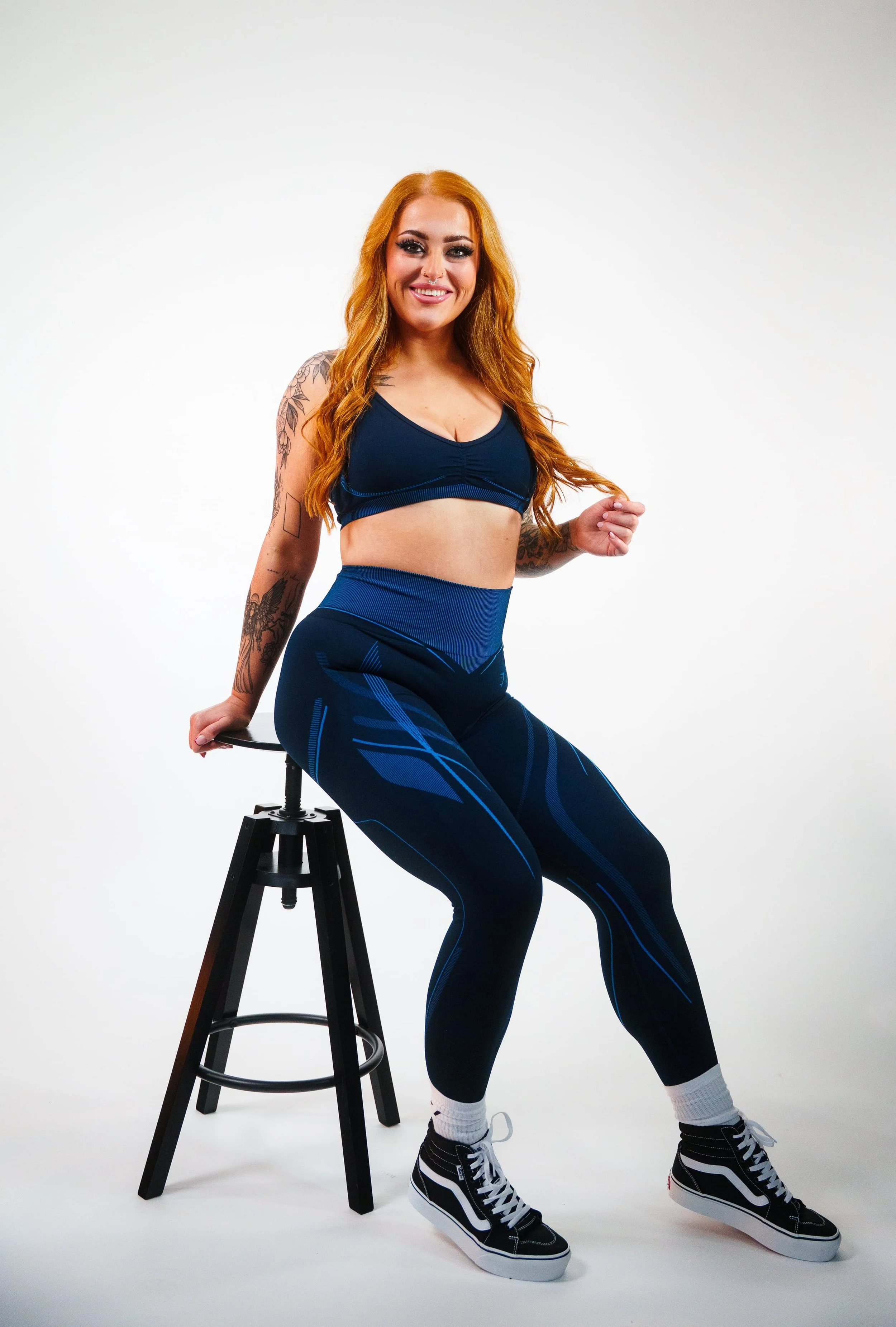 A woman with long red hair, wearing a blue sports bra and matching leggings, sitting on a black stool with one hand resting on it, smiling against a plain white background.