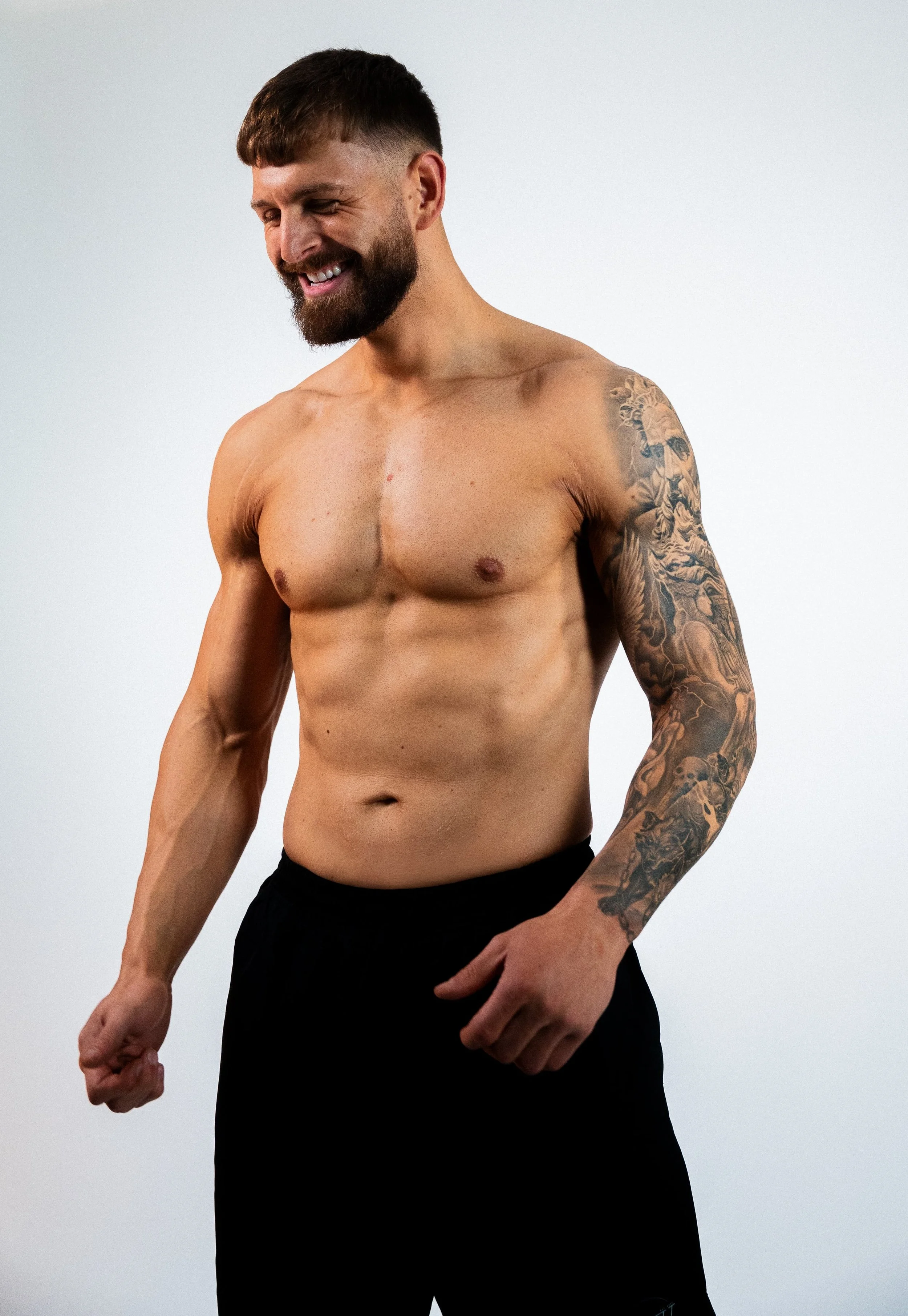 A shirtless man with a beard and tattoos on his left arm, smiling and looking down, wearing black shorts, against a plain light background.