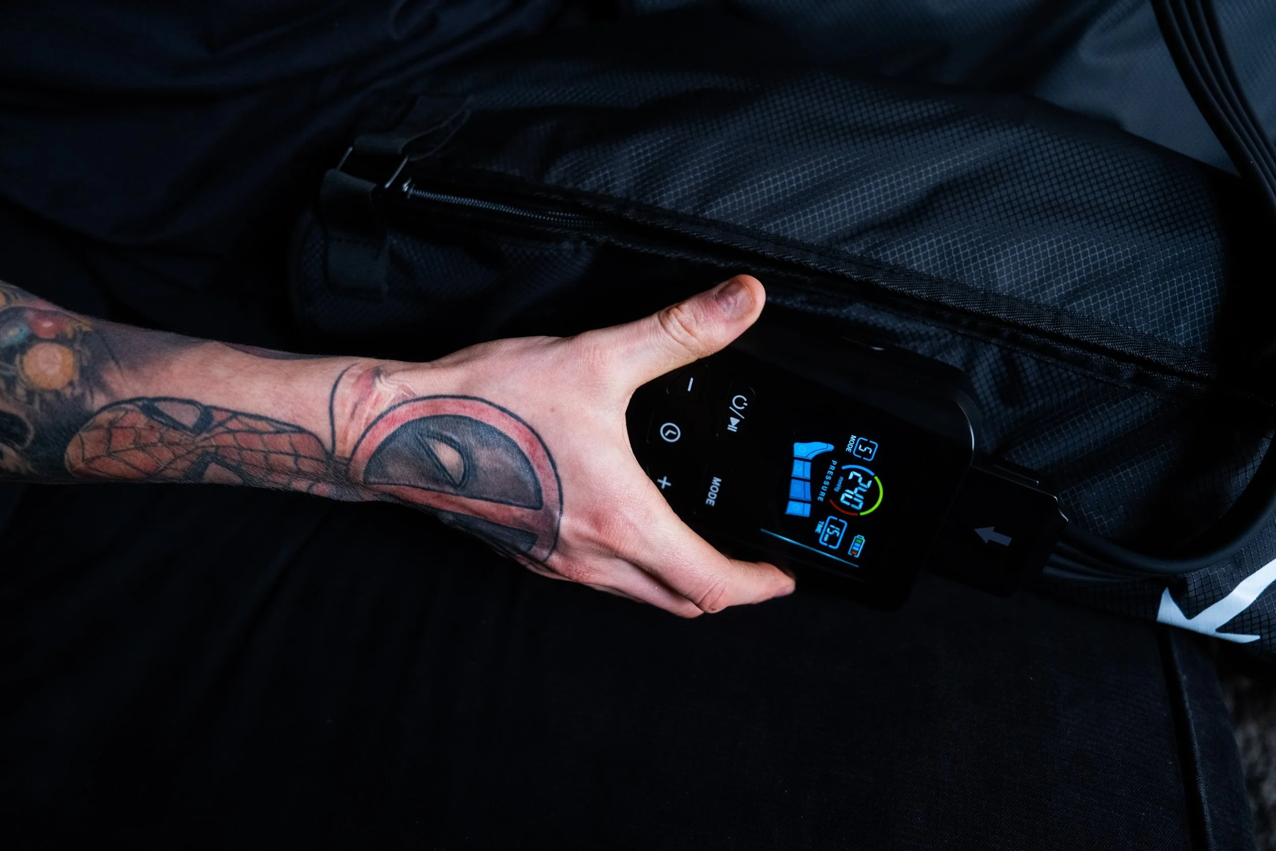 A person with a tattooed arm adjusting a black electronic device with a digital display, connected to a bag, which appears to be a portable pump or control unit.