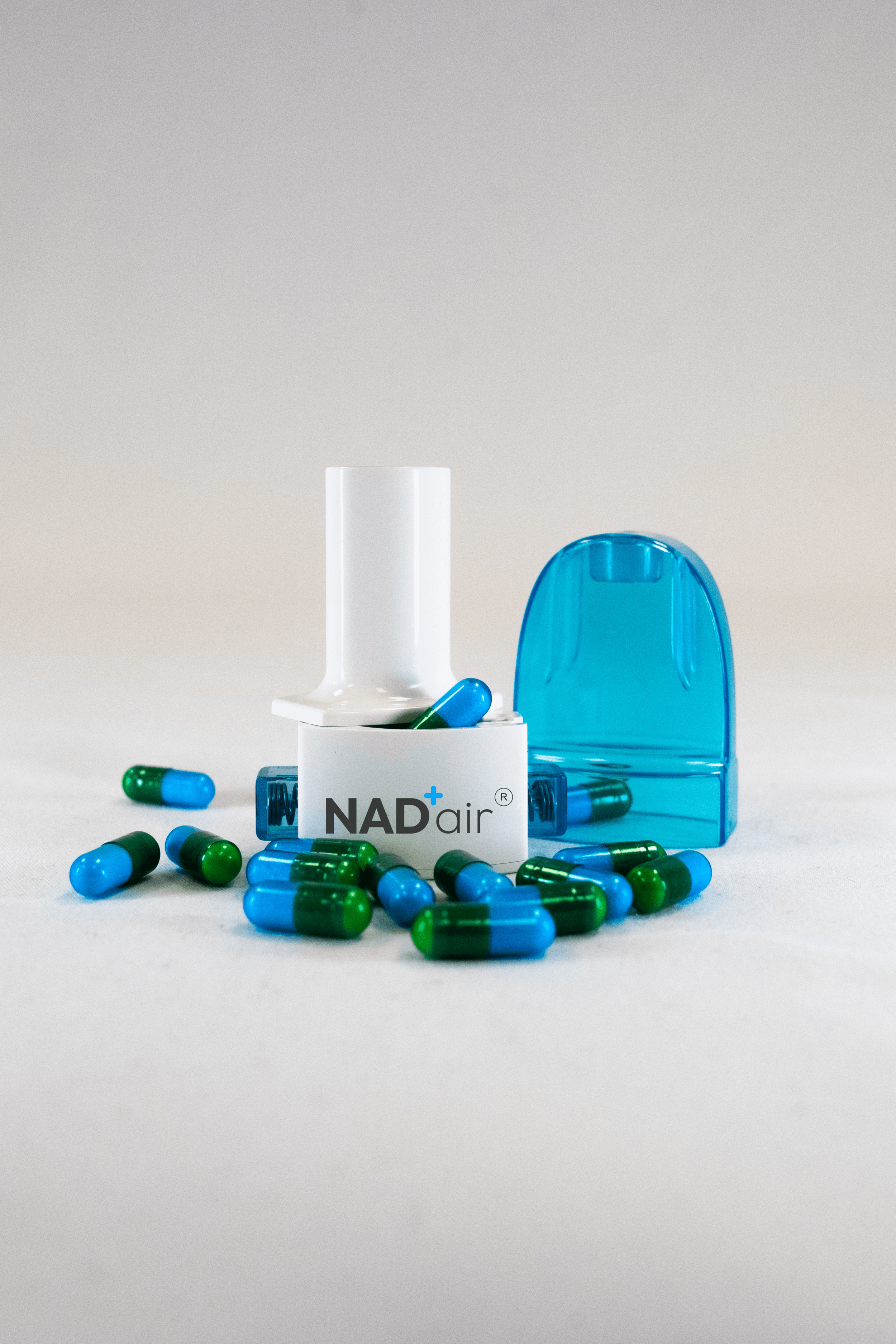 Blue and green capsules scattered around a white medical inhaler labeled 'NAD+ air' with a blue plastic cover on a white surface against a plain background.