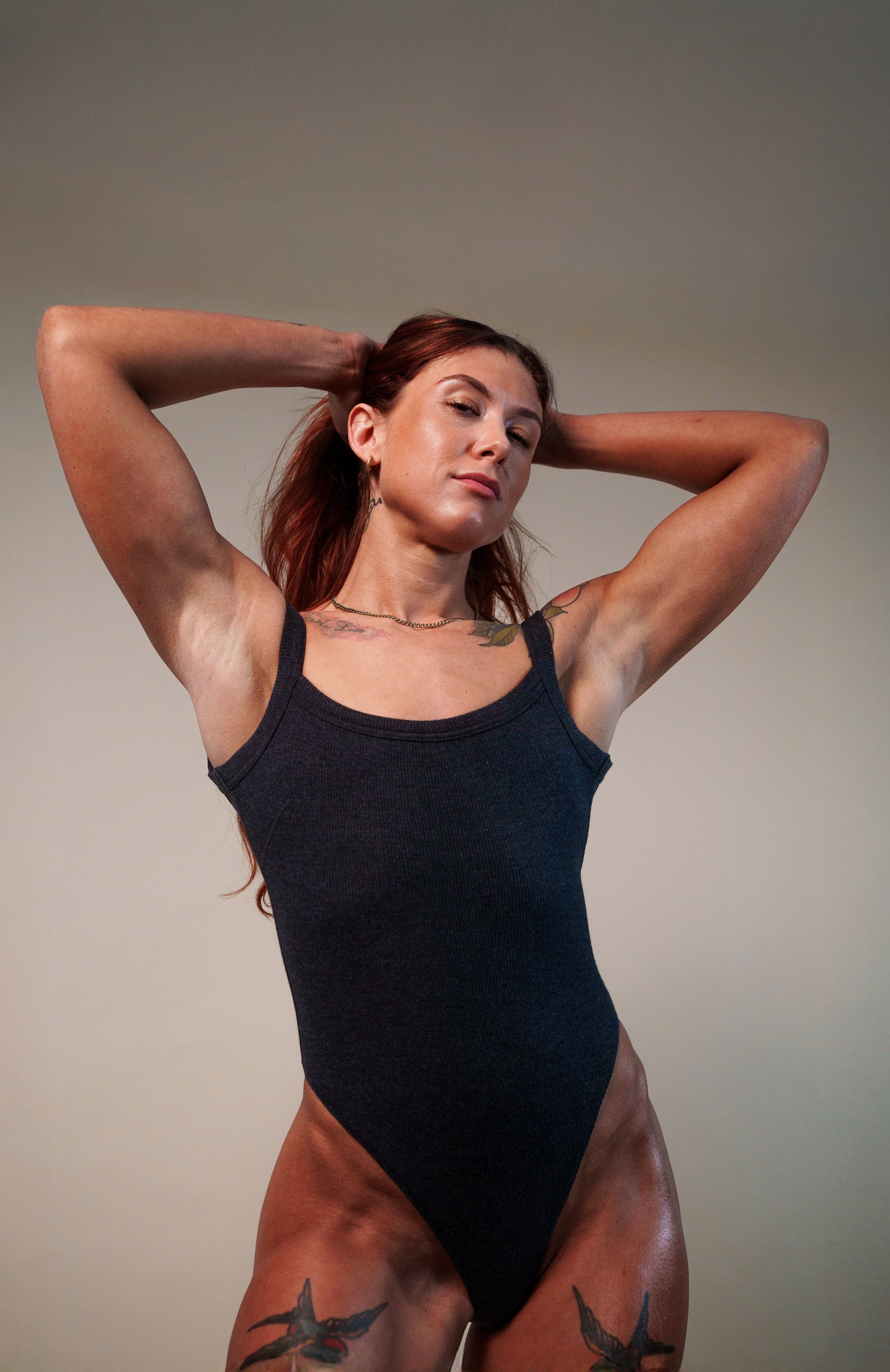 A woman with tattoos wearing a black sleeveless bodysuit, standing against a plain background, with her hands behind her head.