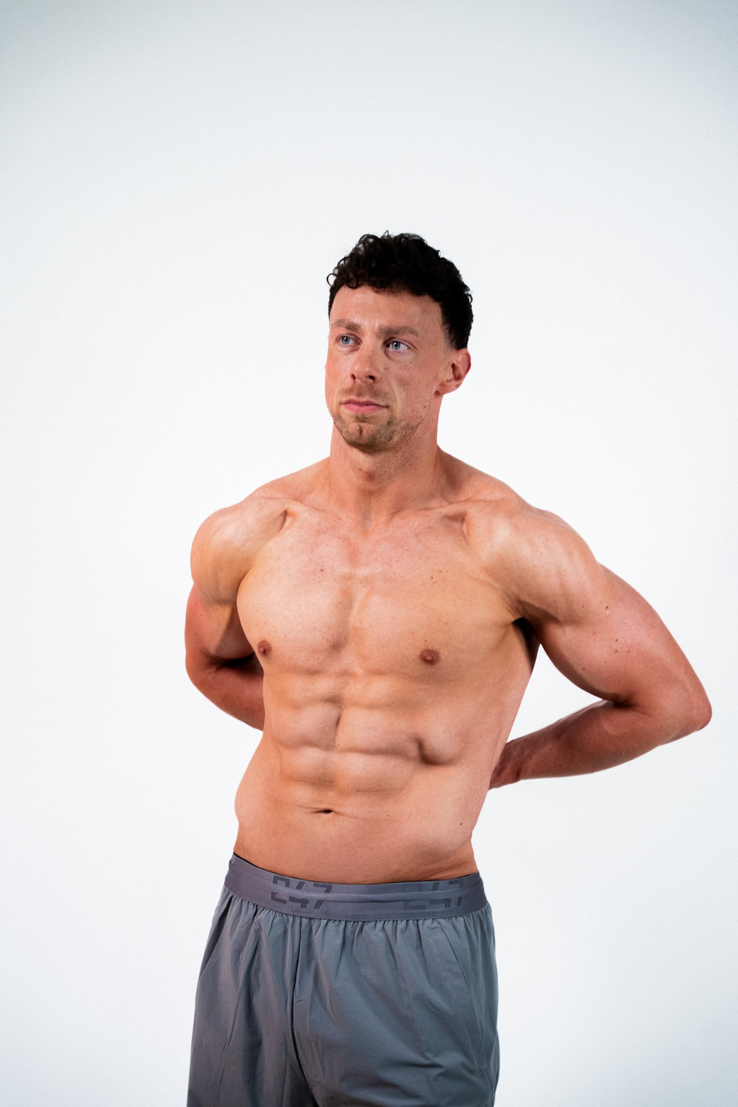 A shirtless man with short dark hair, standing against a plain white background, with his hands behind his back, looking to the side.