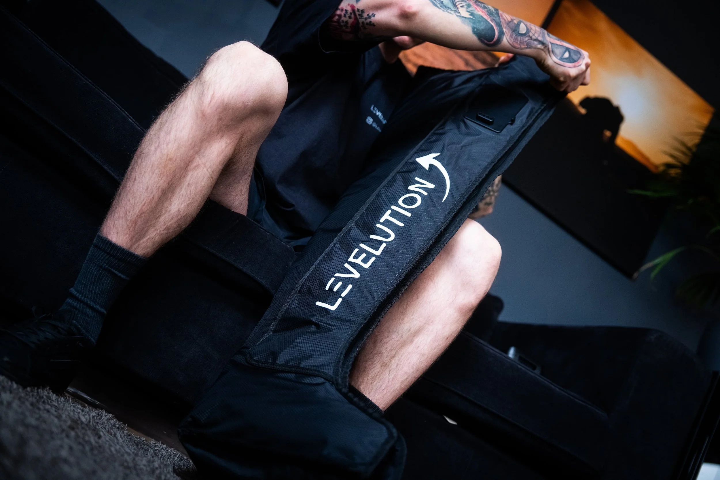 Person sitting on a black couch holding a black equipment bag with the logo 'Levelrevision' in white, in a dimly lit room with a sunset picture on the wall.